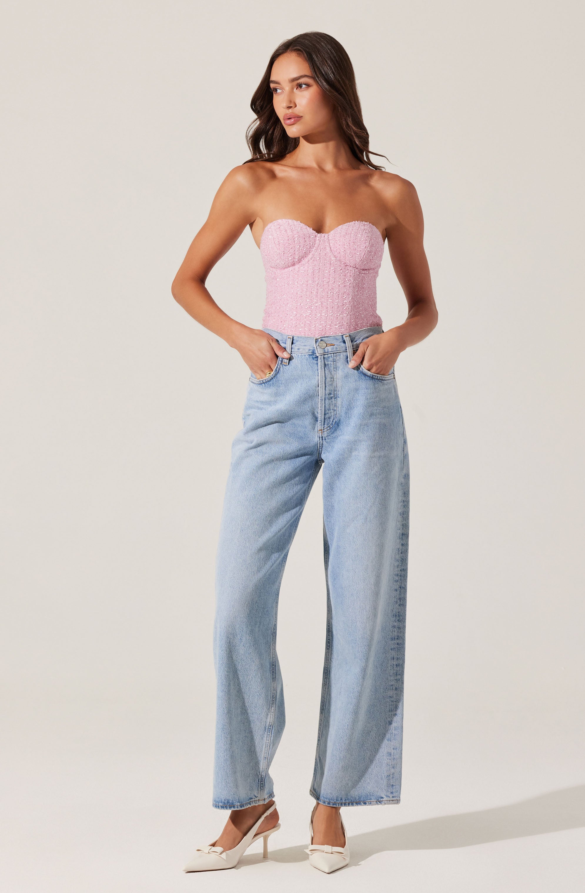 A woman is wearing a strapless pink textured top with a sweetheart neckline and fitted bodice, paired with high-waisted, light-wash wide-leg jeans. She completes the outfit with white pointed-toe heels.