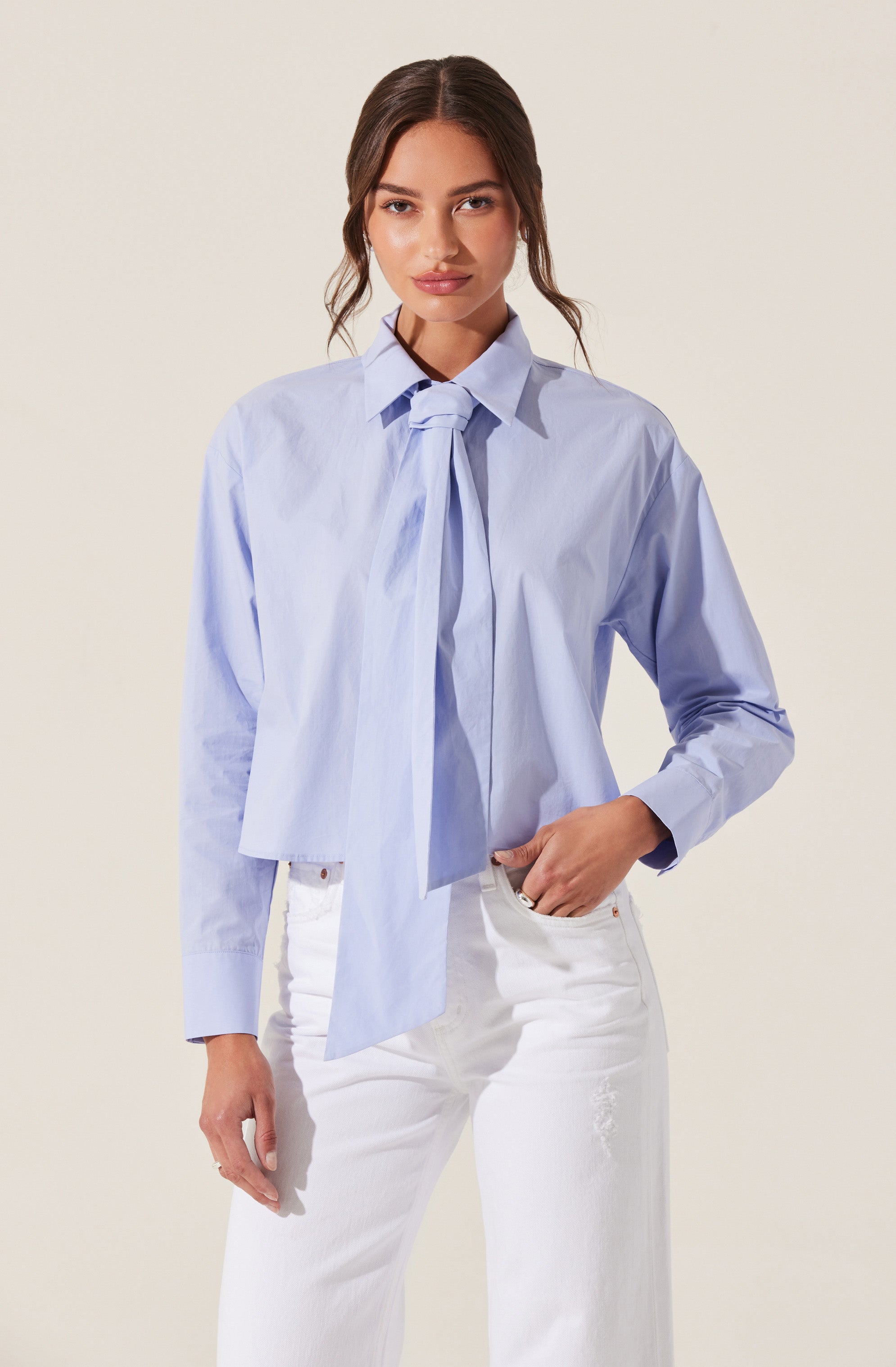 A woman is wearing a light blue button-up shirt with a tie detail at the collar. The shirt has long sleeves and appears slightly oversized. She pairs it with white jeans. Her hair is styled in loose waves, and she has a neutral facial expression.