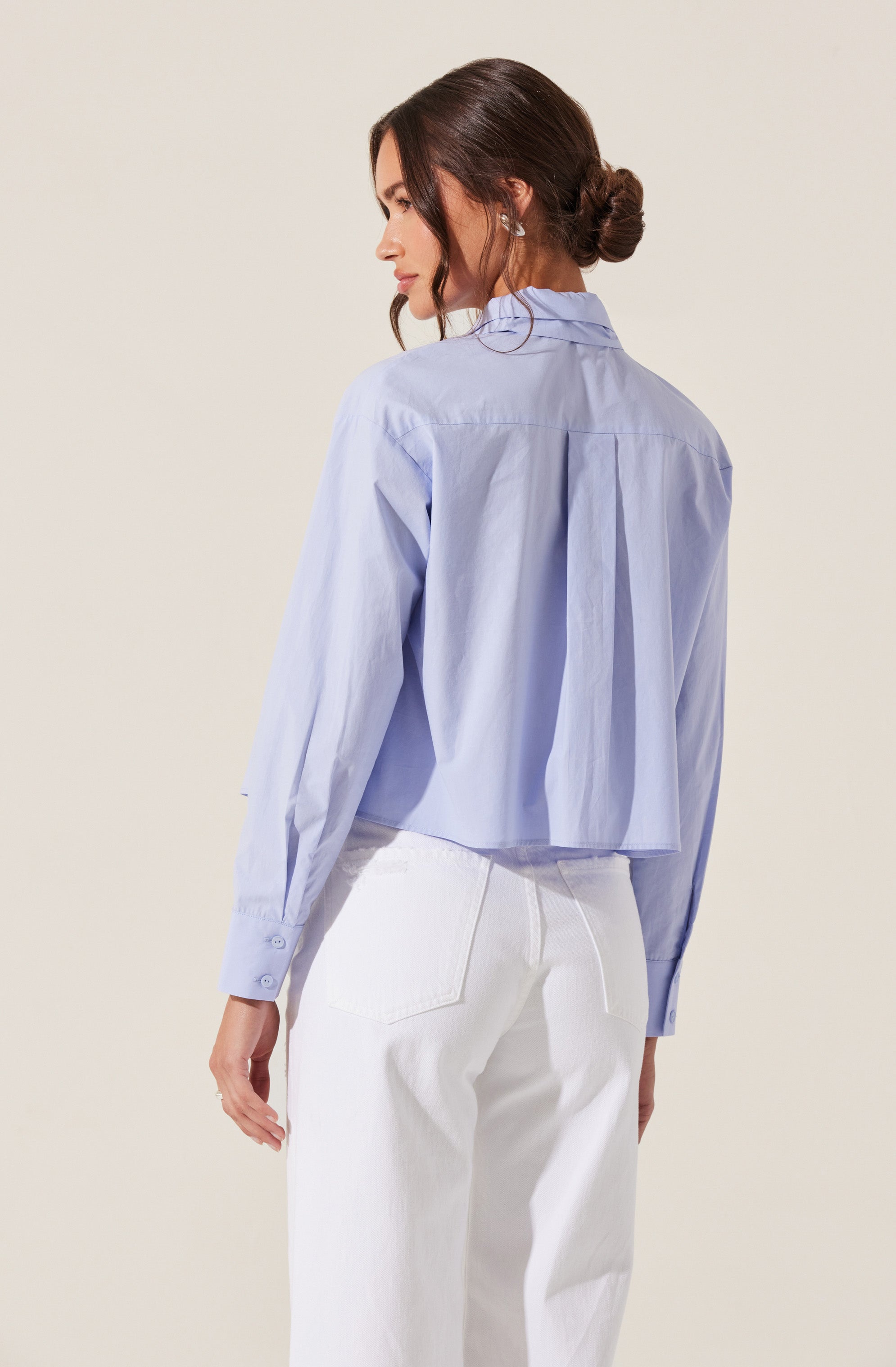 A woman is wearing a light blue, long-sleeved, button-up shirt with a collar and a back pleat detail. She has paired it with high-waisted, white denim jeans.