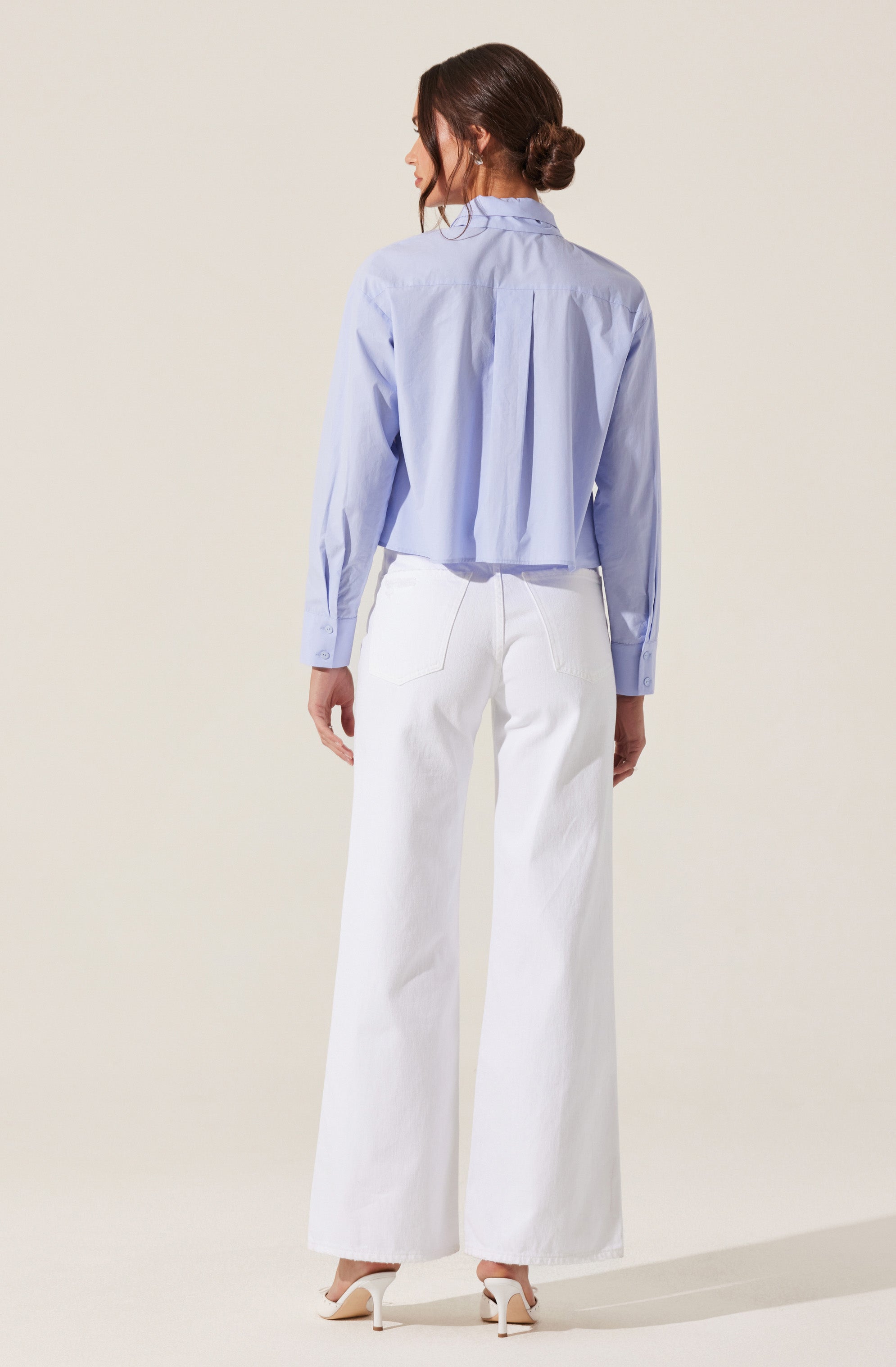 A woman is wearing a light blue long-sleeved button-up shirt with a collar and pleats at the back. She is also wearing high-waisted, wide-leg white pants and beige high-heeled shoes. Her hair is styled in a loose bun.