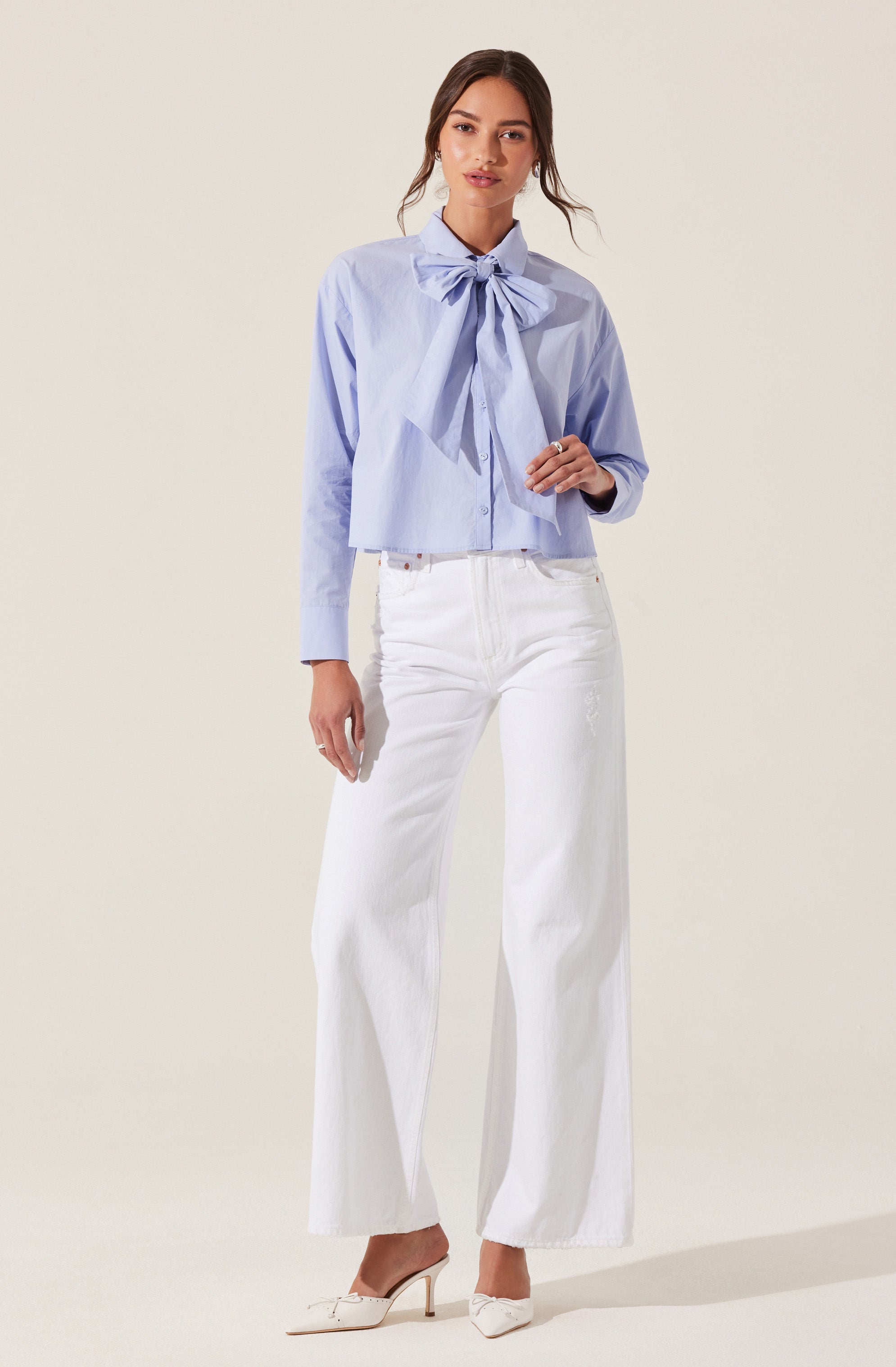 A woman is wearing a light blue blouse with a large bow tie at the collar, paired with high-waisted white wide-leg jeans and white pointed-toe high heels.
