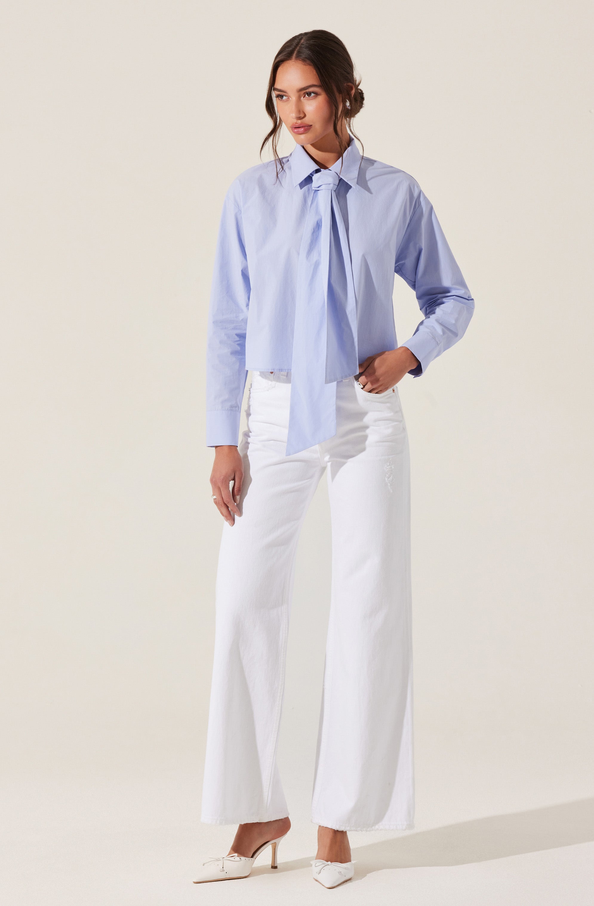 A woman is wearing a light blue button-up shirt with a large, loose tie at the collar. The shirt has long sleeves and a slightly oversized fit. She pairs it with high-waisted, wide-leg white pants and white high-heeled shoes.