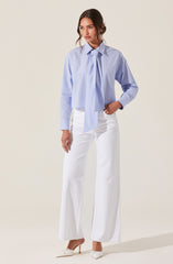 A woman is wearing a light blue button-up shirt with a large, loose tie at the collar. The shirt has long sleeves and a slightly oversized fit. She pairs it with high-waisted, wide-leg white pants and white high-heeled shoes.