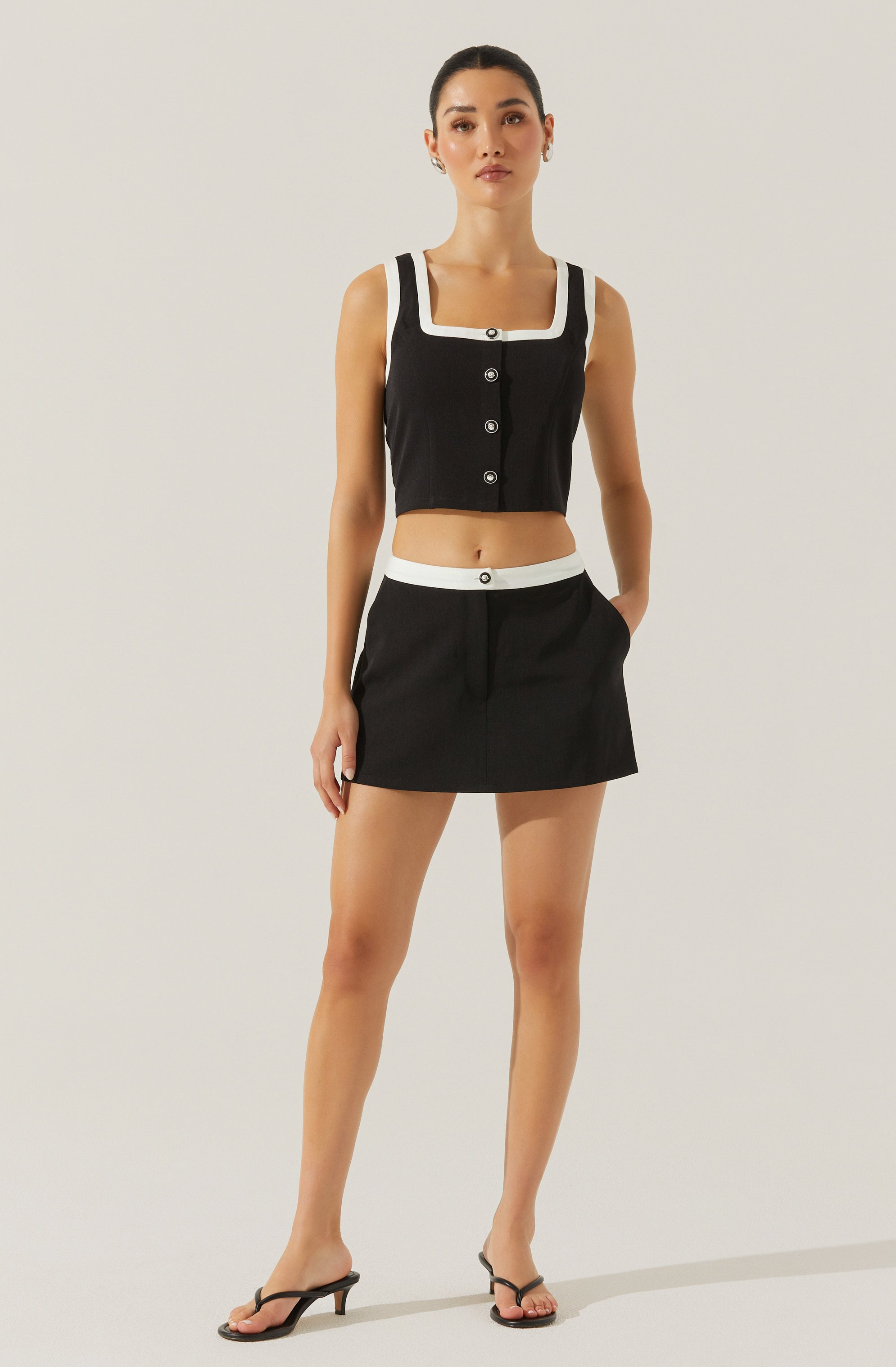 A woman is wearing a black sleeveless crop top with white trim along the square neckline and armholes, featuring black buttons down the front. She pairs the top with a matching black mini skirt that has a white waistband and white button detail at the center. She completes her outfit with black open-toe high-heeled sandals.