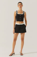 A woman is wearing a black sleeveless crop top with white trim along the square neckline and armholes, featuring black buttons down the front. She pairs the top with a matching black mini skirt that has a white waistband and white button detail at the center. She completes her outfit with black open-toe high-heeled sandals.