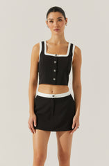 A woman is wearing a black sleeveless cropped top with white trim around the neckline, sleeve edges, and button placket, paired with a matching black mini skirt that has a white waistband with a single black button.