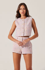 A young woman is wearing a sleeveless, pastel-colored, textured tweed vest with a round neckline and a row of buttons down the front. She pairs it with matching high-waisted shorts that have a similar pastel, textured tweed fabric, and a front zipper. The fabric has a multicolored appearance with light pink, blue, and cream tones.