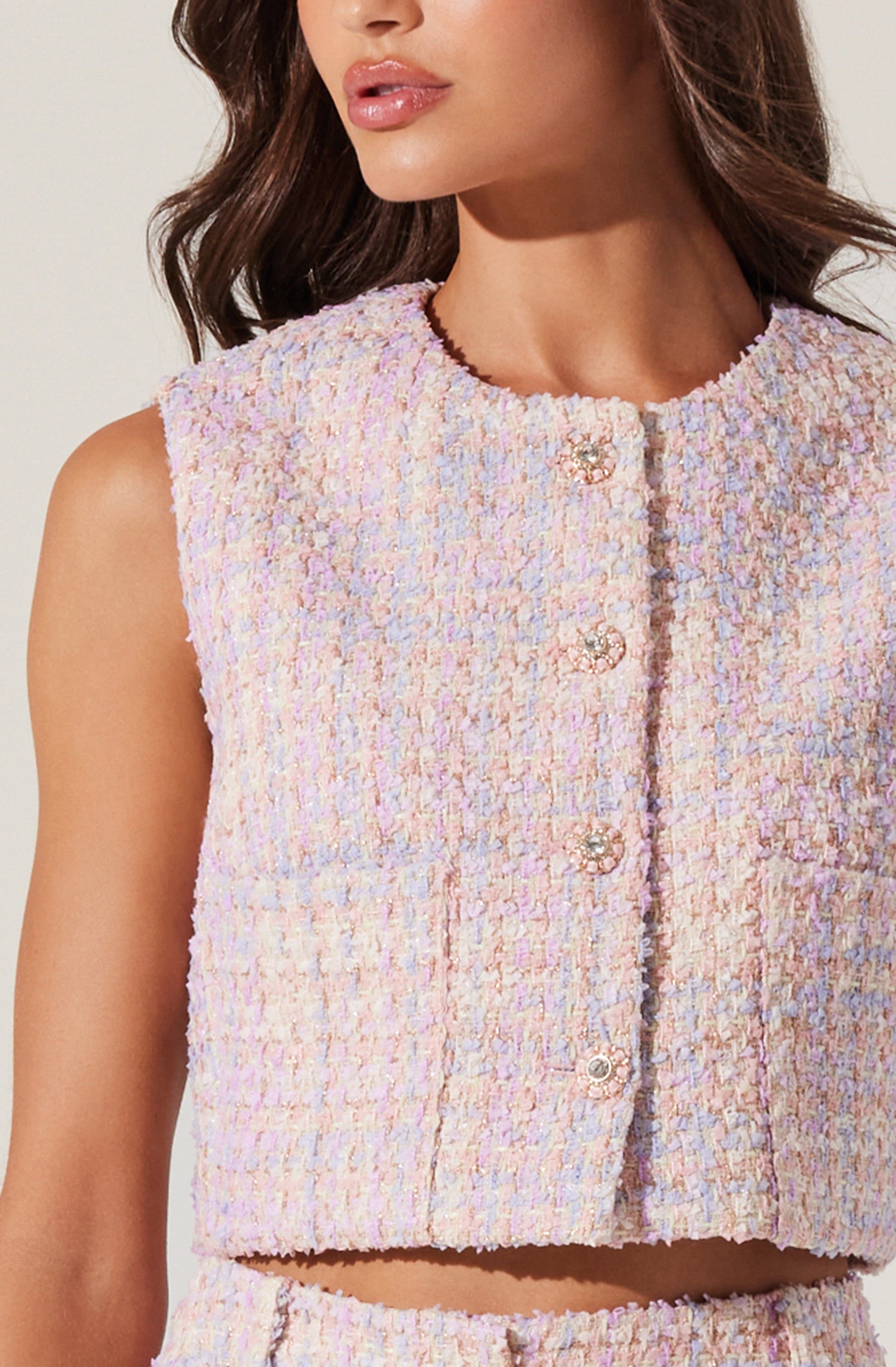 A sleeveless, cropped top made of textured tweed fabric in pastel shades of pink, lavender, and cream. The top features a round neckline and a front closure with decorative pearl and metal button details.