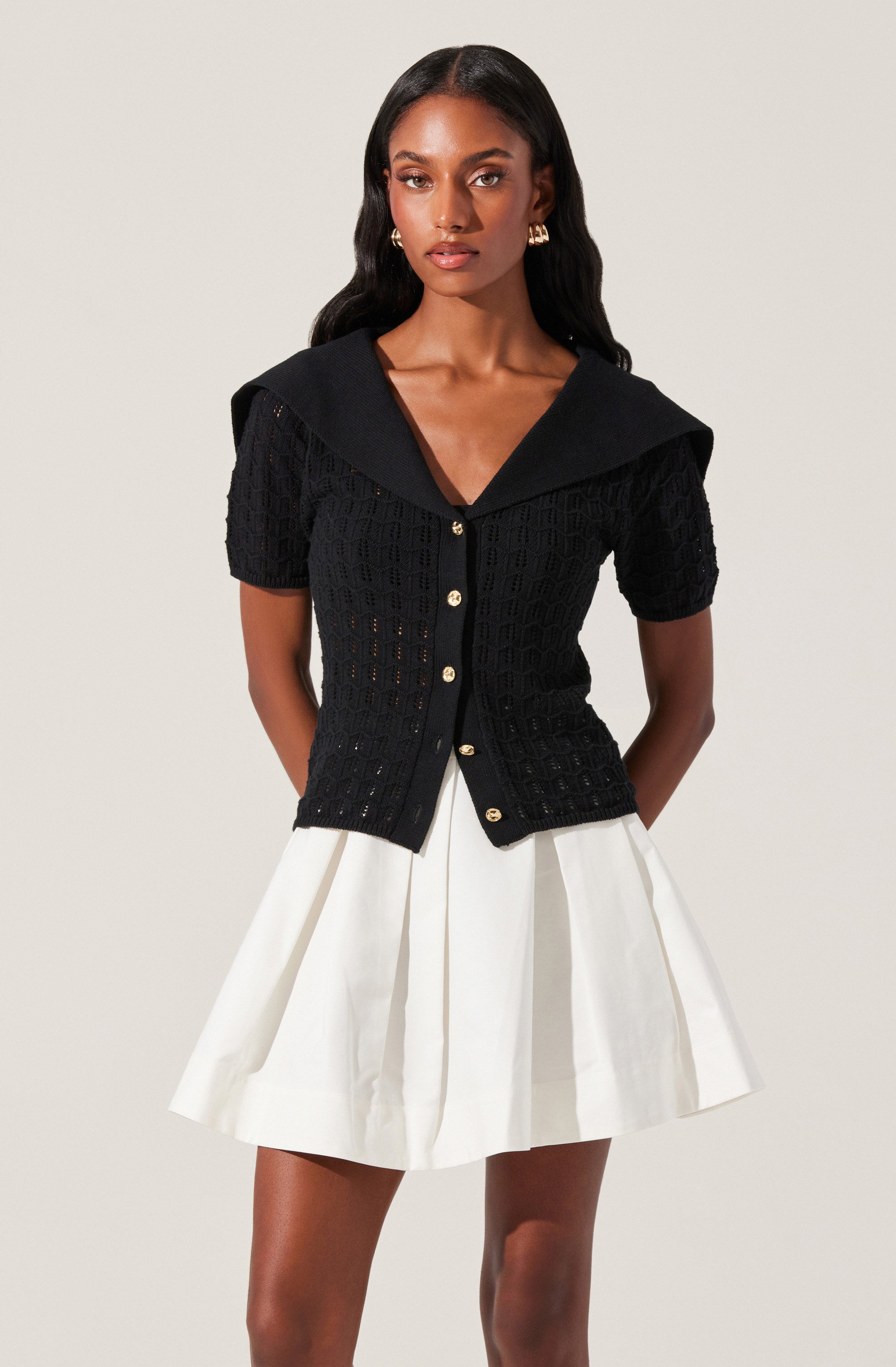 The woman is wearing a black short-sleeved knit cardigan with a large collar that drapes over her shoulders and gold buttons down the front. She pairs it with a white, pleated, A-line skirt.