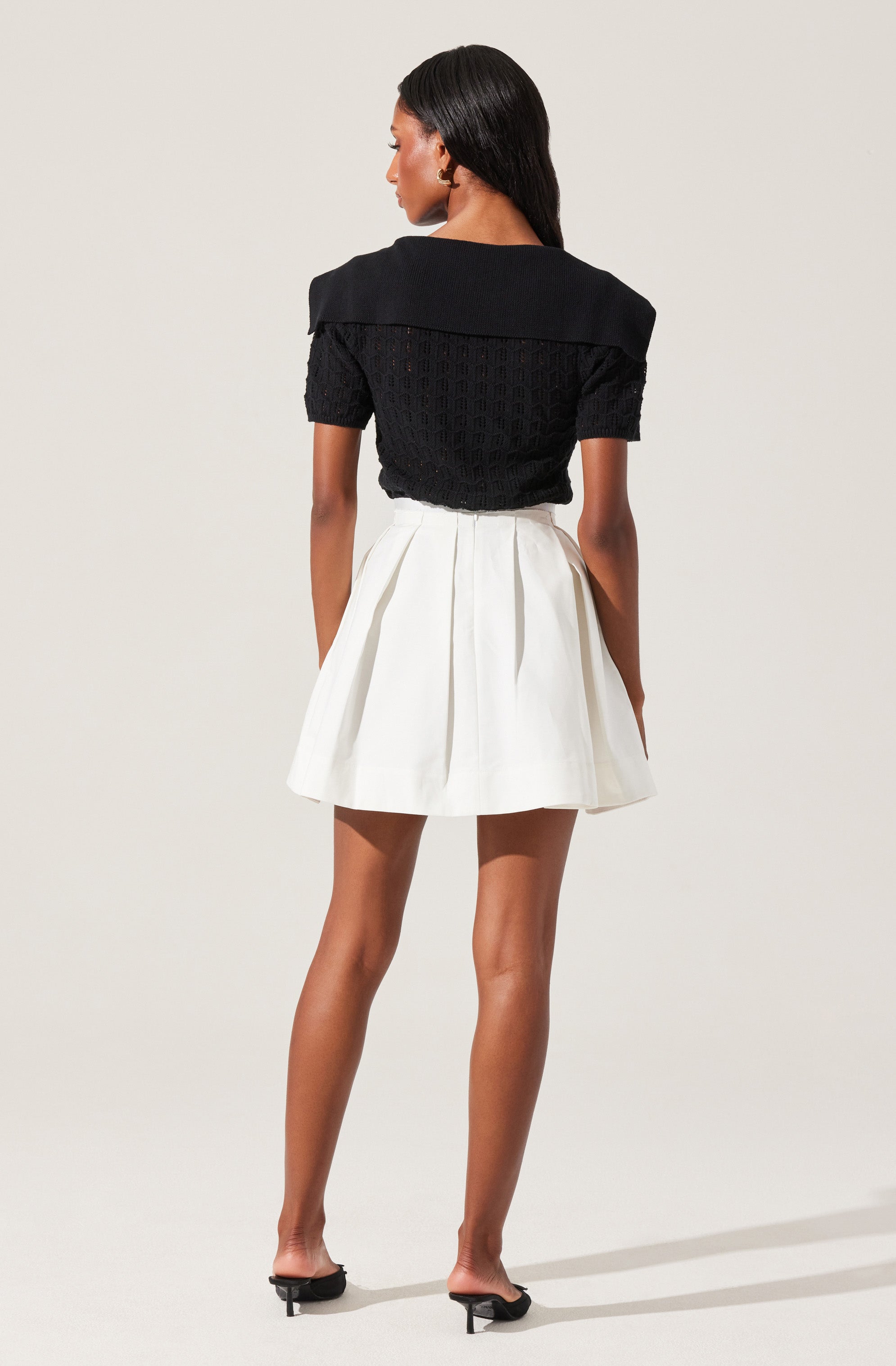 A woman is wearing a black short-sleeved top with a wide collar and a textured knit pattern. She has paired it with a high-waisted white pleated skirt. She is also wearing black open-toe heels.