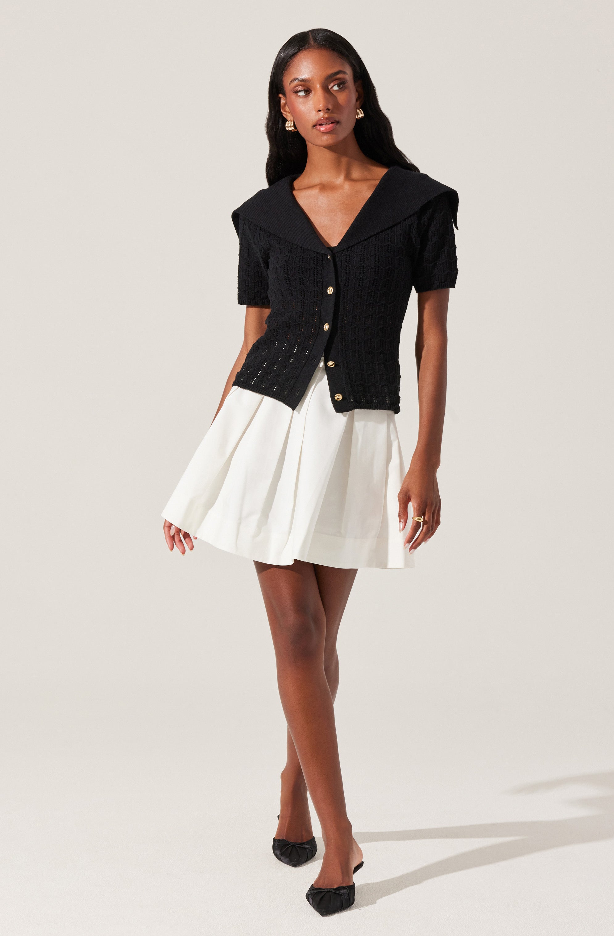 A woman is wearing a short-sleeved black knit cardigan with a large collar and gold buttons down the front. She pairs it with a white pleated skirt and black pointed-toe heels.