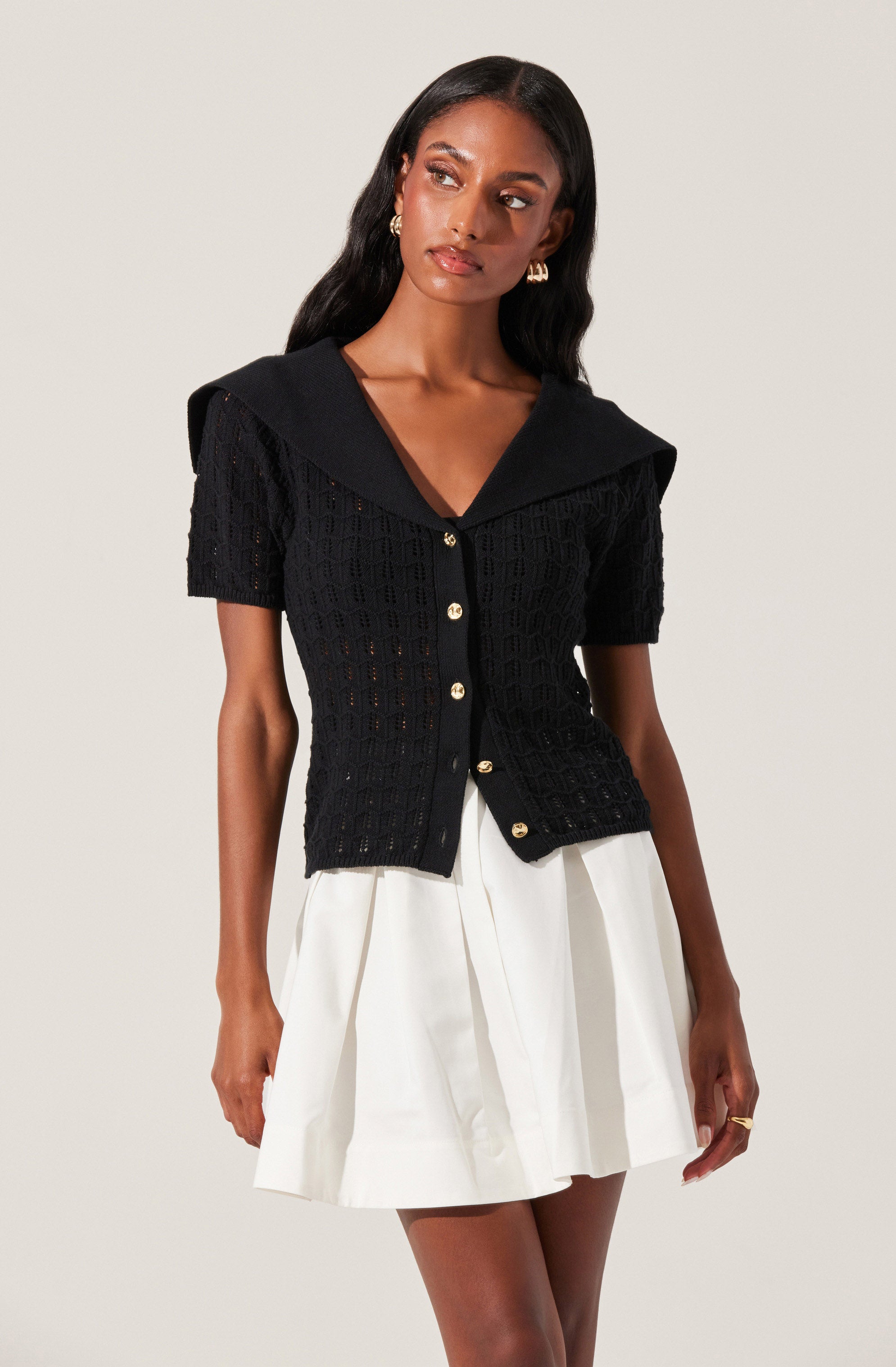 A woman wearing a black, short-sleeved, open-knit top with a wide collar and gold buttons down the front. The top has a textured, crochet-like pattern. She pairs it with a high-waisted, white, pleated skirt. She accessorizes with gold earrings and a ring.