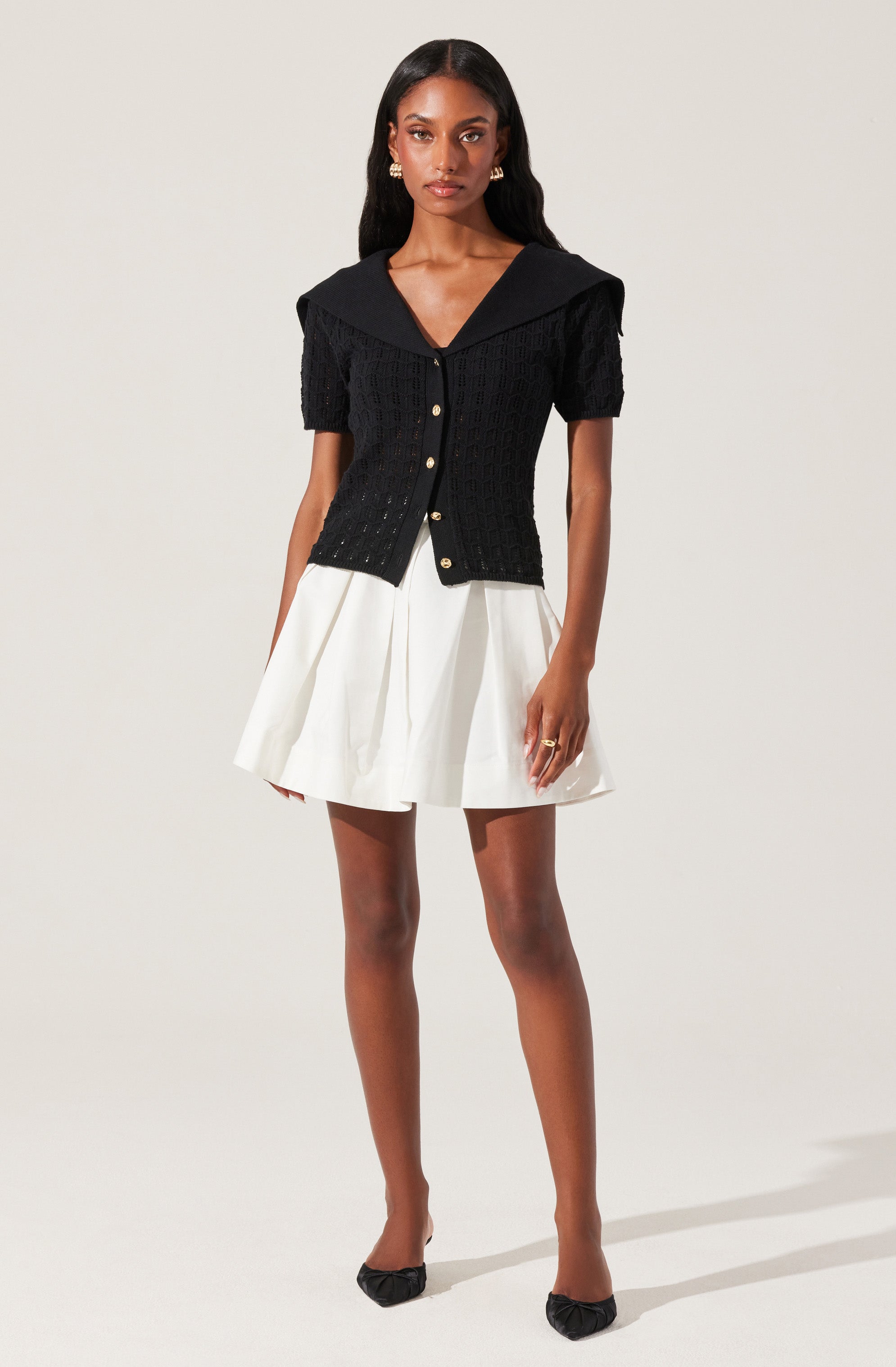 A woman is wearing a black short-sleeved knitted cardigan with pearl buttons down the front, paired with a white pleated skirt that reaches above the knees. She has accessorized with gold earrings and a ring, and is wearing black high-heeled shoes.