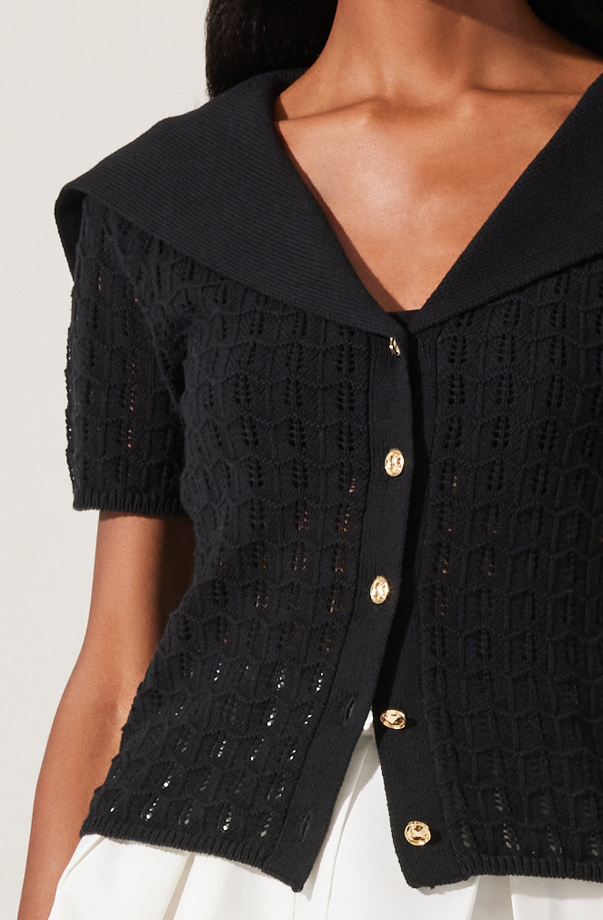 A person is wearing a black, short-sleeved, open-knit cardigan with a textured geometric pattern. The cardigan features a wide collar and gold buttons down the front. They are also wearing white pants.
