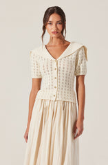 A woman is wearing a cream-colored short-sleeved knitted top with a V-neckline and a wide collar that drapes over her shoulders. The top features a square-shaped open-knit pattern and is buttoned down the front with small, round, light-colored buttons. She is also wearing a long, flowing skirt in a matching light cream or beige shade with pleats.