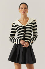 A woman is wearing a long-sleeved, black and white striped cardigan with gold buttons down the front and on the cuffs. The cardigan features a wide, white, sailor-style collar. She pairs it with a high-waisted, black pleated skirt. She has gold hoop earrings and a gold ring on her finger.