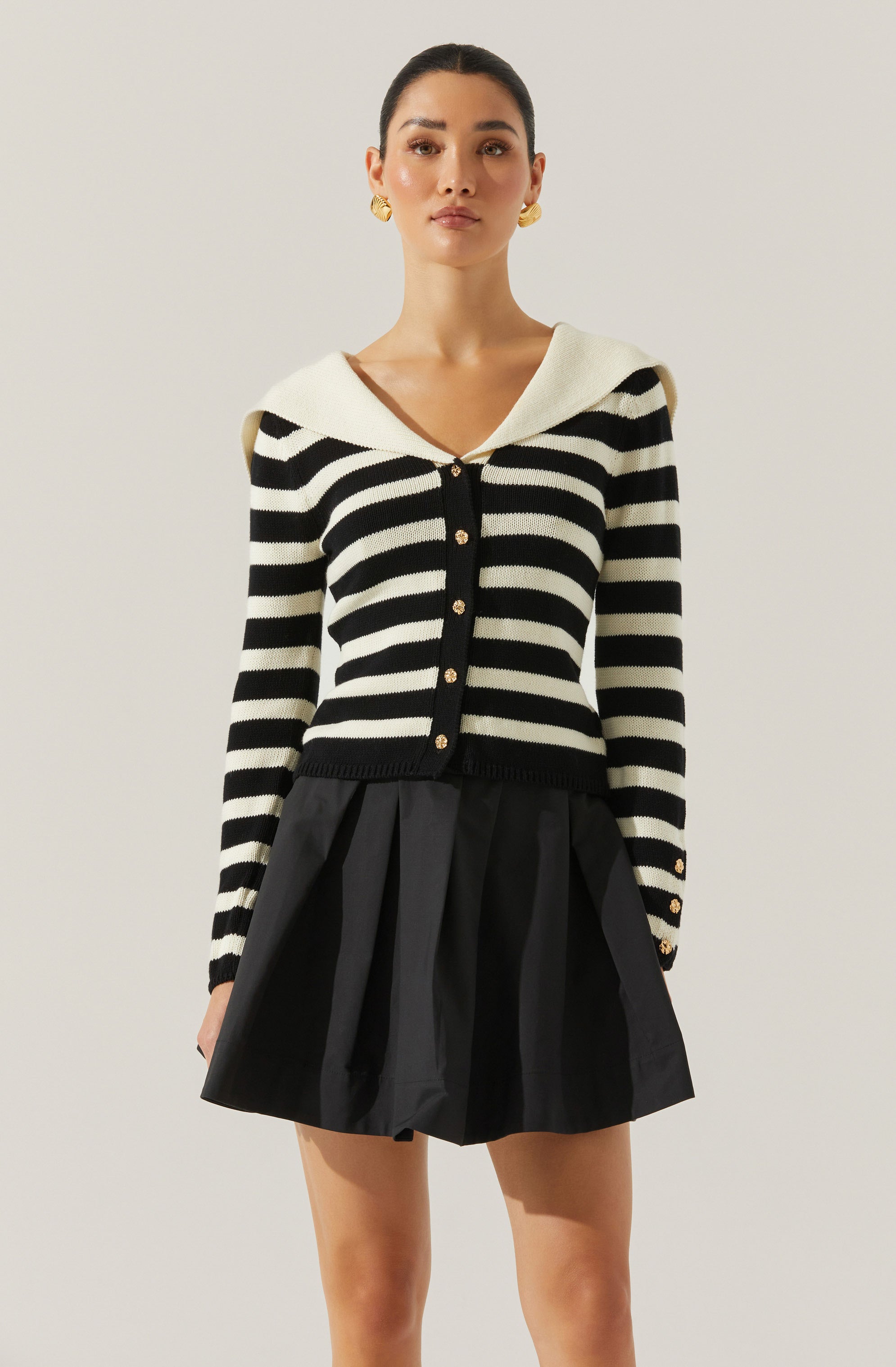 A woman is wearing a long-sleeve, black and white striped cardigan with a wide, white, folded collar. The cardigan features a row of gold buttons down the front and on the cuffs. She is also wearing a black skirt with pleats.