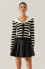 A woman is wearing a long-sleeve, black and white striped cardigan with a wide, white, folded collar. The cardigan features a row of gold buttons down the front and on the cuffs. She is also wearing a black skirt with pleats.