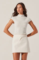 A woman wearing a sleeveless, high-neck white knitted top paired with a white skirt featuring a small slit on the left side and a thin belt with a buckle.
