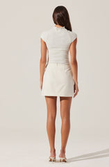 A woman is wearing a fitted short-sleeve, light gray top with a high neckline, paired with a high-waisted, cream-colored mini skirt that has a slightly flared silhouette and subtle pleats. She completes the look with white heeled shoes.