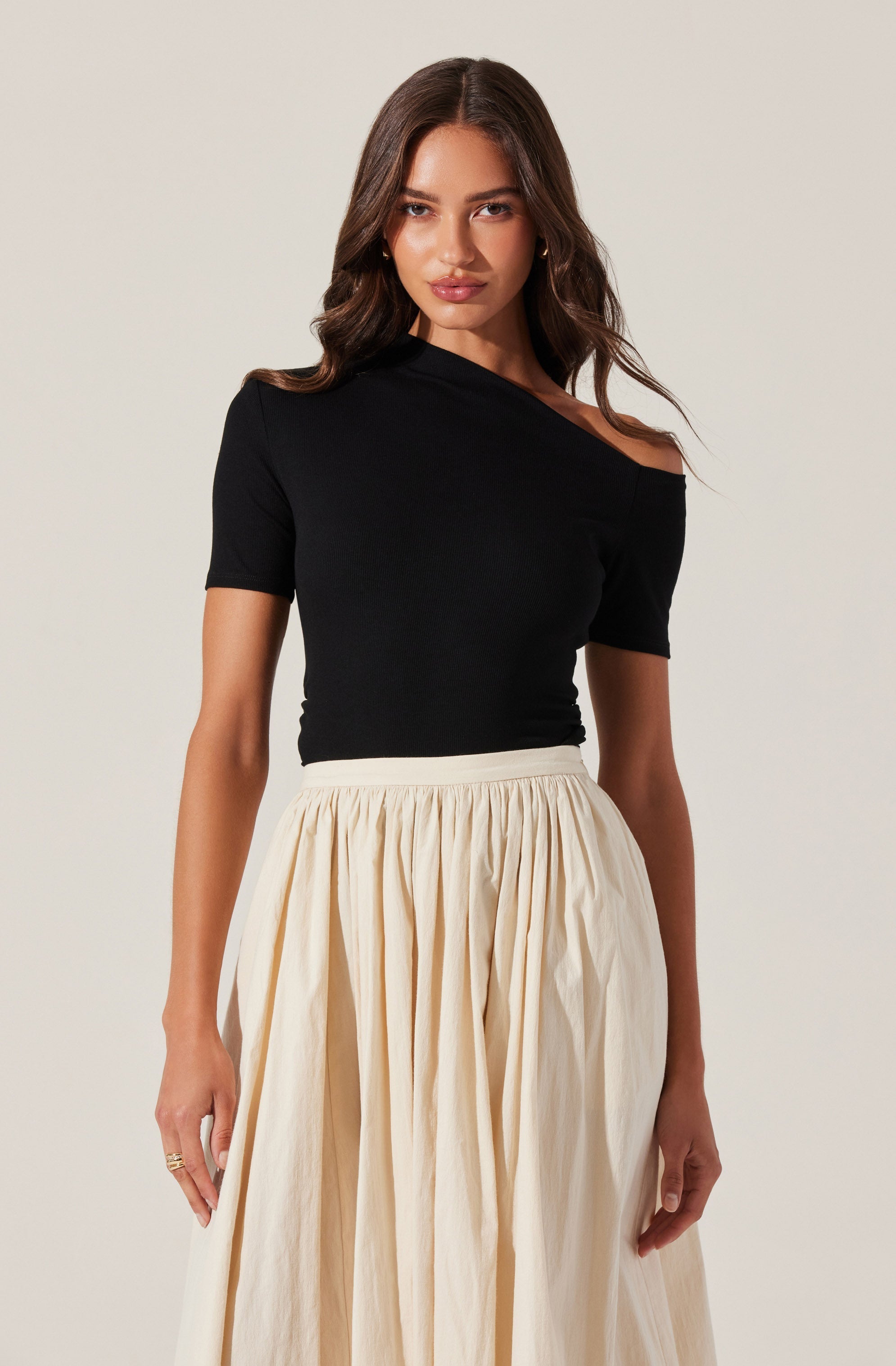 A woman is wearing a black, off-the-shoulder top with short sleeves, paired with a high-waisted, cream-colored, pleated skirt that reaches below the knees.