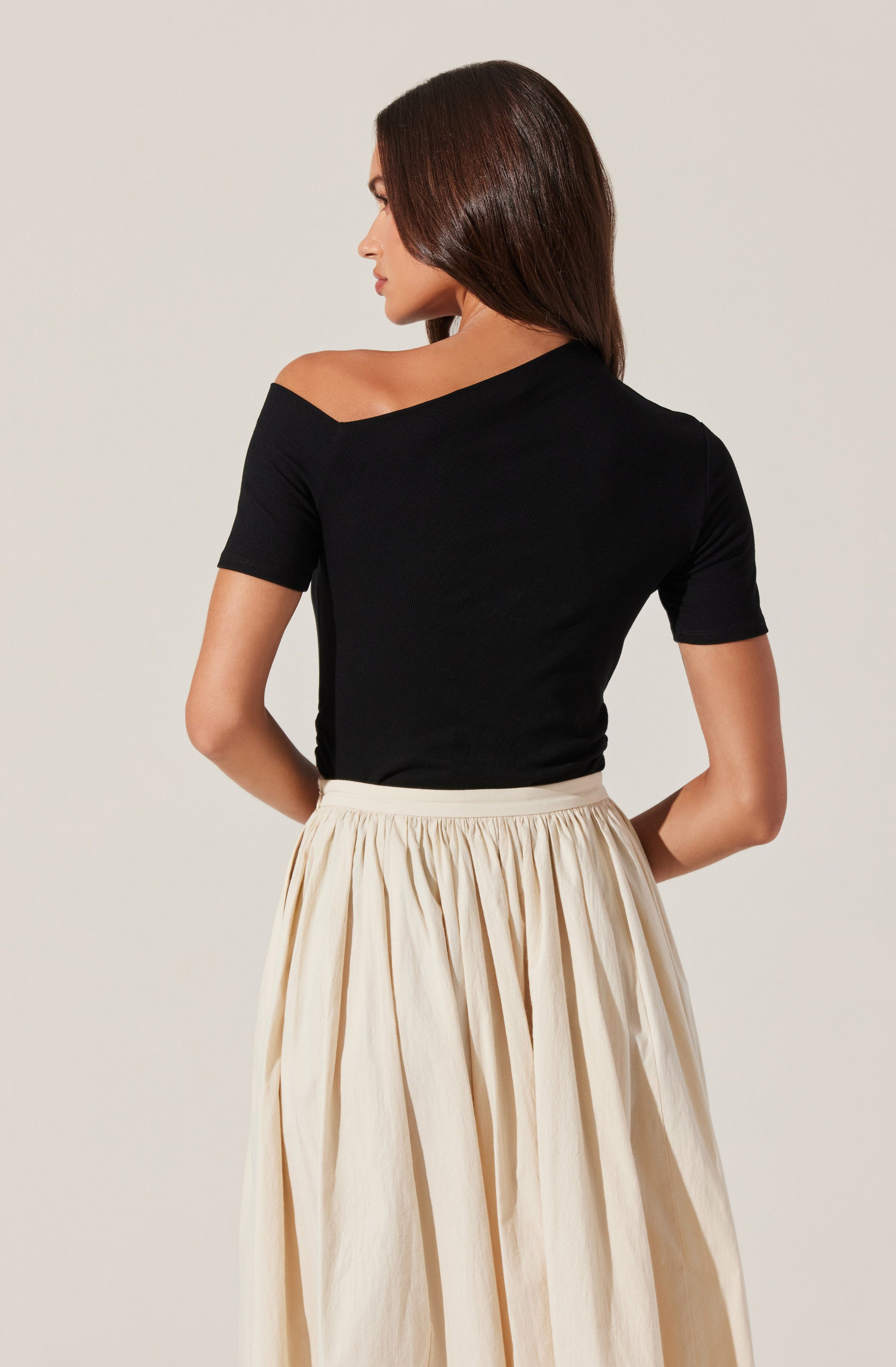A woman is wearing an off-the-shoulder black top with short sleeves, paired with a high-waisted, light-colored, gathered skirt.