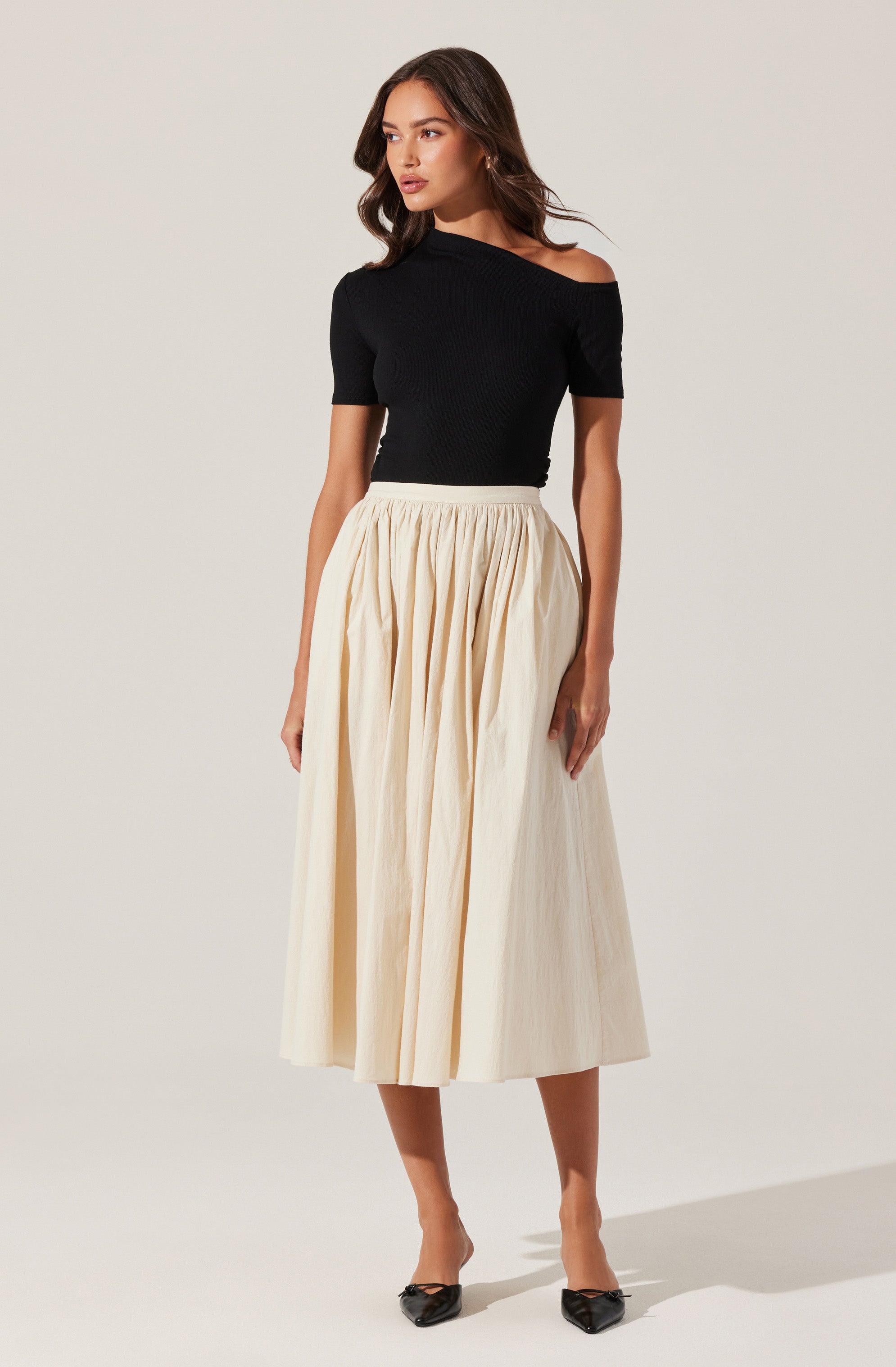 A woman is wearing a black off-the-shoulder short-sleeve top paired with a high-waisted, full-length cream-colored pleated skirt. She is also wearing black pointed-toe heels.