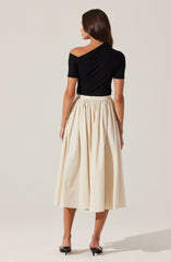 A woman is wearing a black, off-the-shoulder top with short sleeves, paired with a high-waisted, cream-colored, pleated midi skirt. She is also wearing black, open-toe, low-heeled shoes.