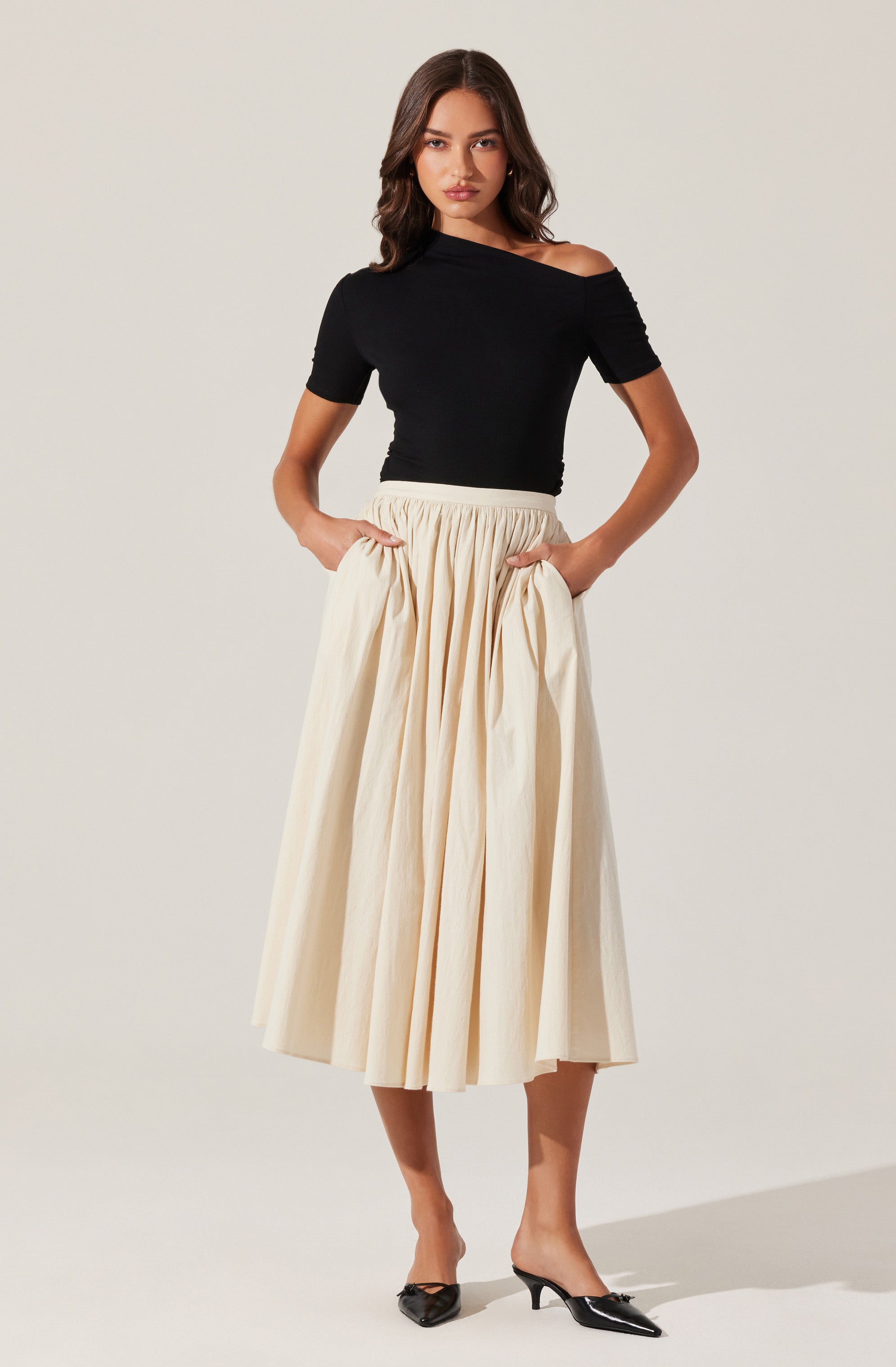 A woman is wearing a black, off-the-shoulder, short-sleeved top and a high-waisted, beige, full-length skirt with pleats. She is also wearing pointed black heels.