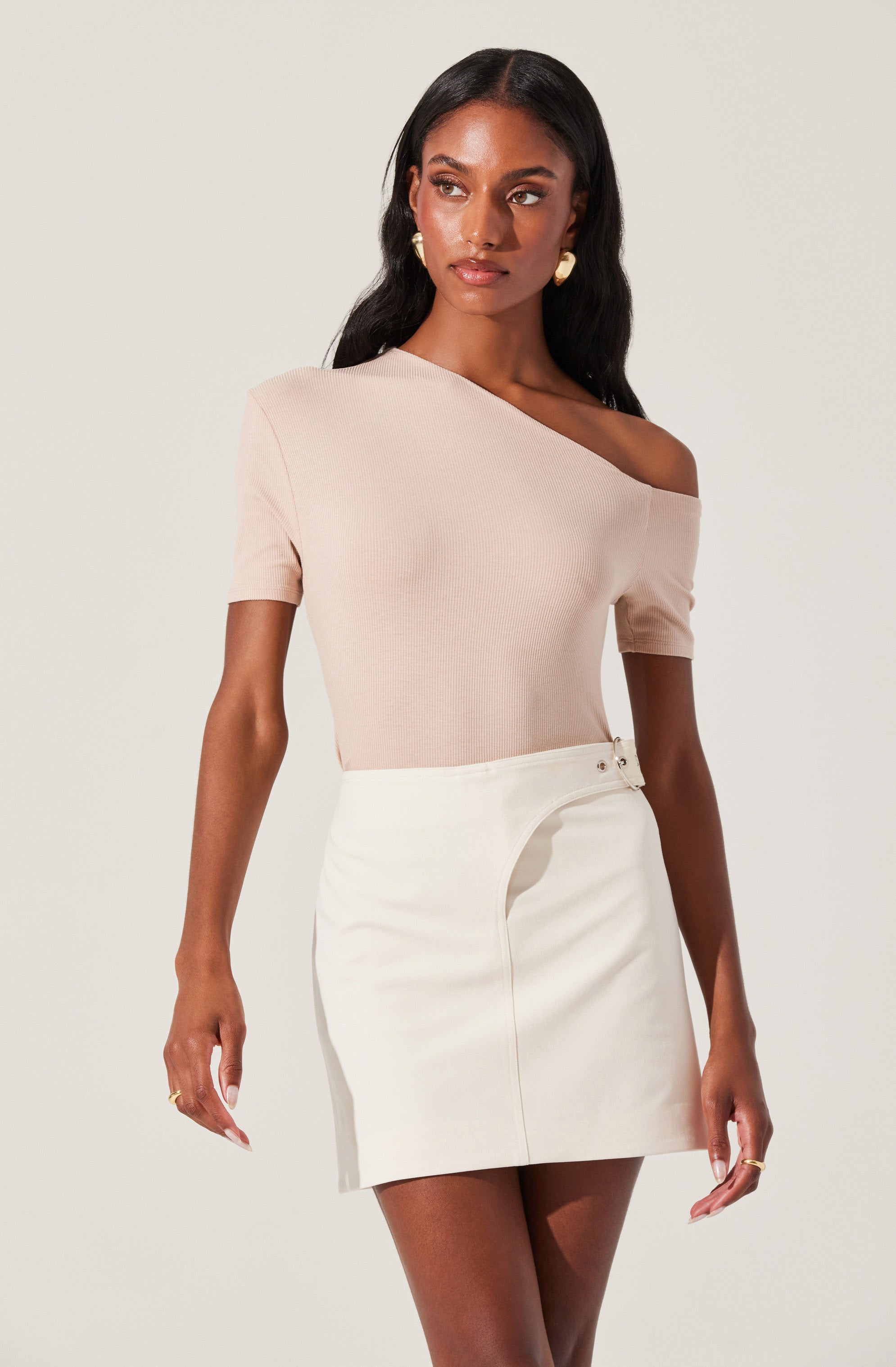 A woman is wearing a light pink, asymmetrical, off-the-shoulder top with short sleeves, paired with a high-waisted, off-white mini skirt that has a subtle slit on the front and a small metal detail on the waistband.