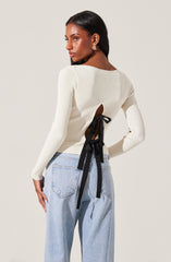 A woman is wearing a cream-colored, long-sleeved, ribbed sweater with a distinctive back design featuring a large cutout tied with two black ribbons. She pairs it with high-waisted, light-wash jeans.