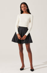 A woman is wearing a long-sleeved, off-white, ribbed sweater with a boat neckline. She pairs it with a high-waisted, black pleated mini skirt. She completes the look with black pointed-toe heels and gold earrings.