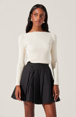 A woman is wearing a fitted, long-sleeved, ribbed sweater in an off-white or cream color. She pairs it with a high-waisted black skirt that has pleats and a flared silhouette. She accessorizes with gold earrings and rings.