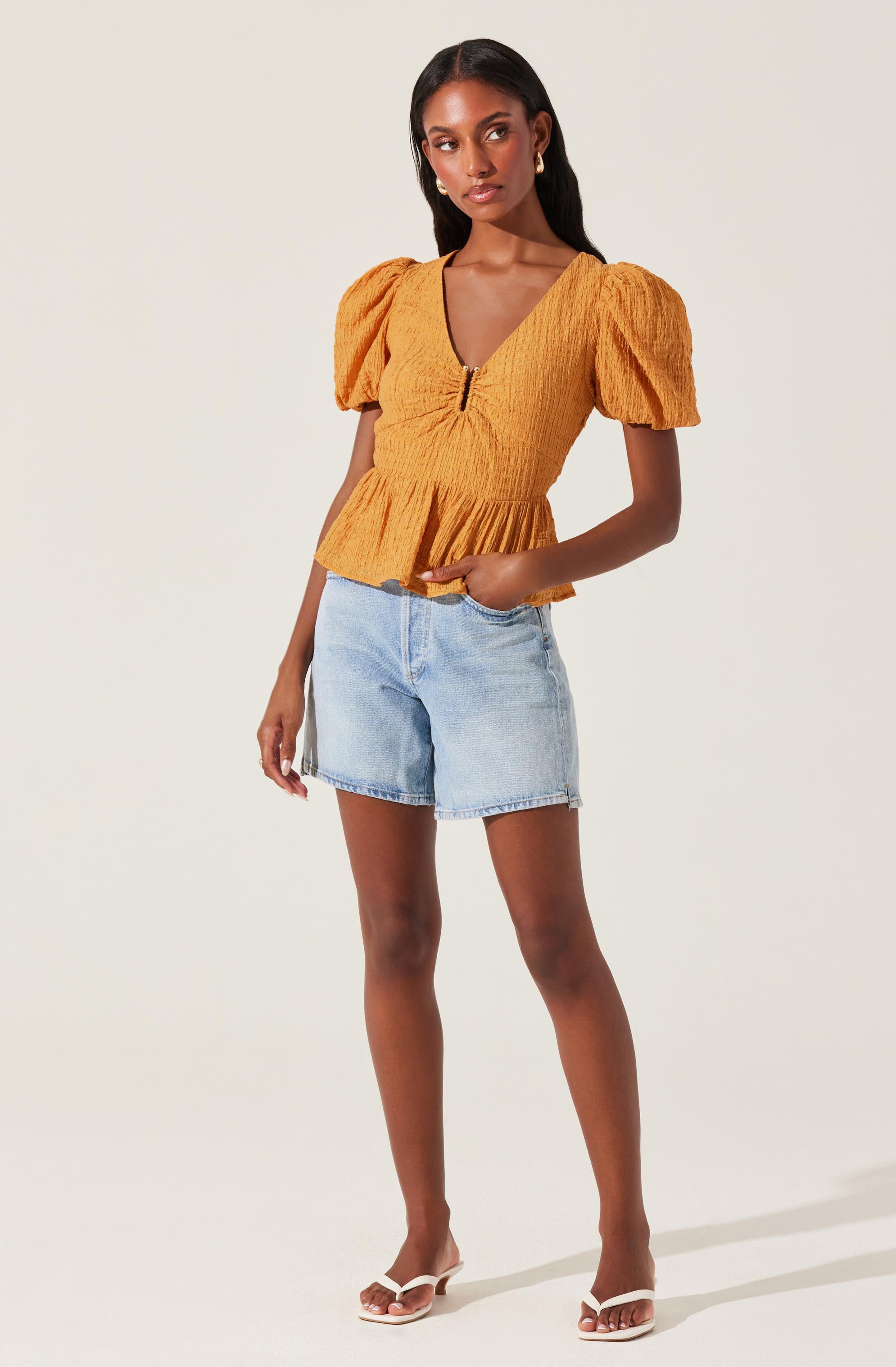 A woman is wearing a mustard yellow top with puffed short sleeves and a fitted, textured bodice featuring a front zipper and a peplum hem. She pairs it with light blue denim shorts and white heeled sandals.