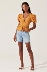 A woman is wearing a mustard yellow top with puffed short sleeves and a fitted, textured bodice featuring a front zipper and a peplum hem. She pairs it with light blue denim shorts and white heeled sandals.