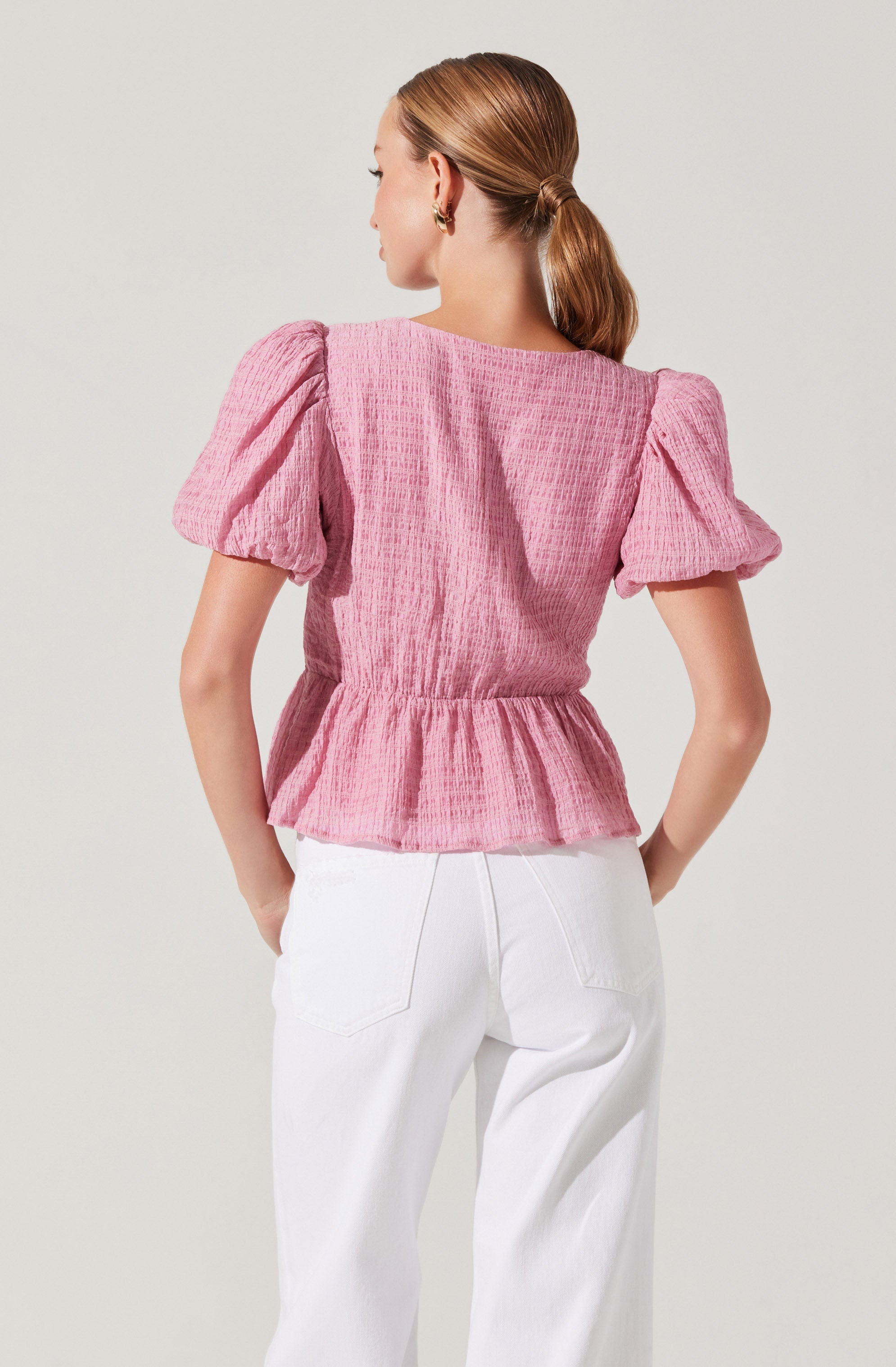 A woman is wearing a pink textured top with puffy short sleeves and a gathered waist, paired with high-waisted white pants. Her hair is styled in a low ponytail, and she has small earrings.