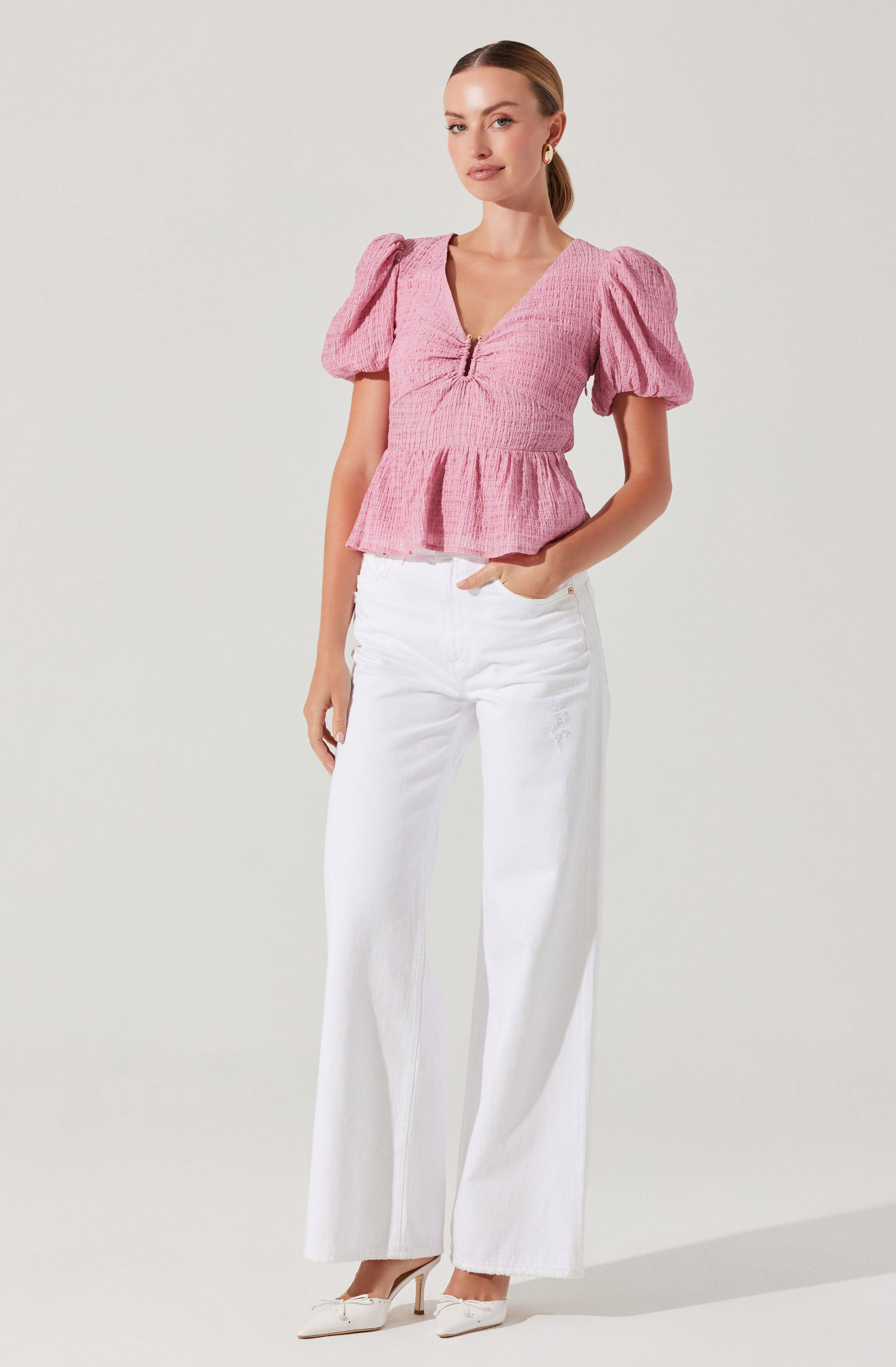 The woman is wearing a pink textured top with puffed short sleeves, a V-neckline, and a front zipper. The top features a gathered detail at the bust and a peplum-style hem. She pairs it with high-waisted, wide-leg white jeans and white pointed toe heels.