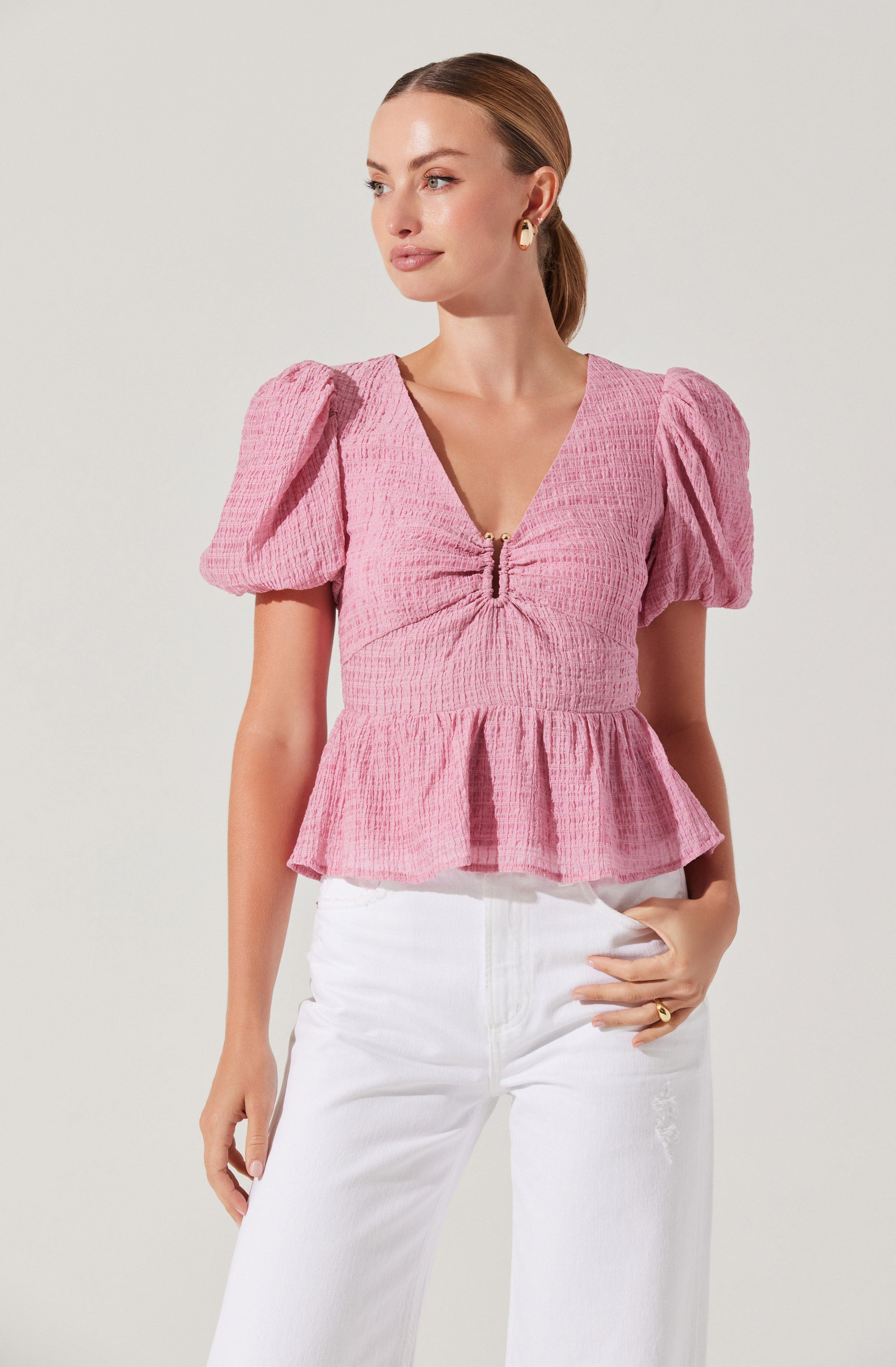 A woman wearing a pink, textured blouse with puffed short sleeves, a V-neckline with a front zipper, and a ruffled hem. She pairs the blouse with white high-waisted pants.