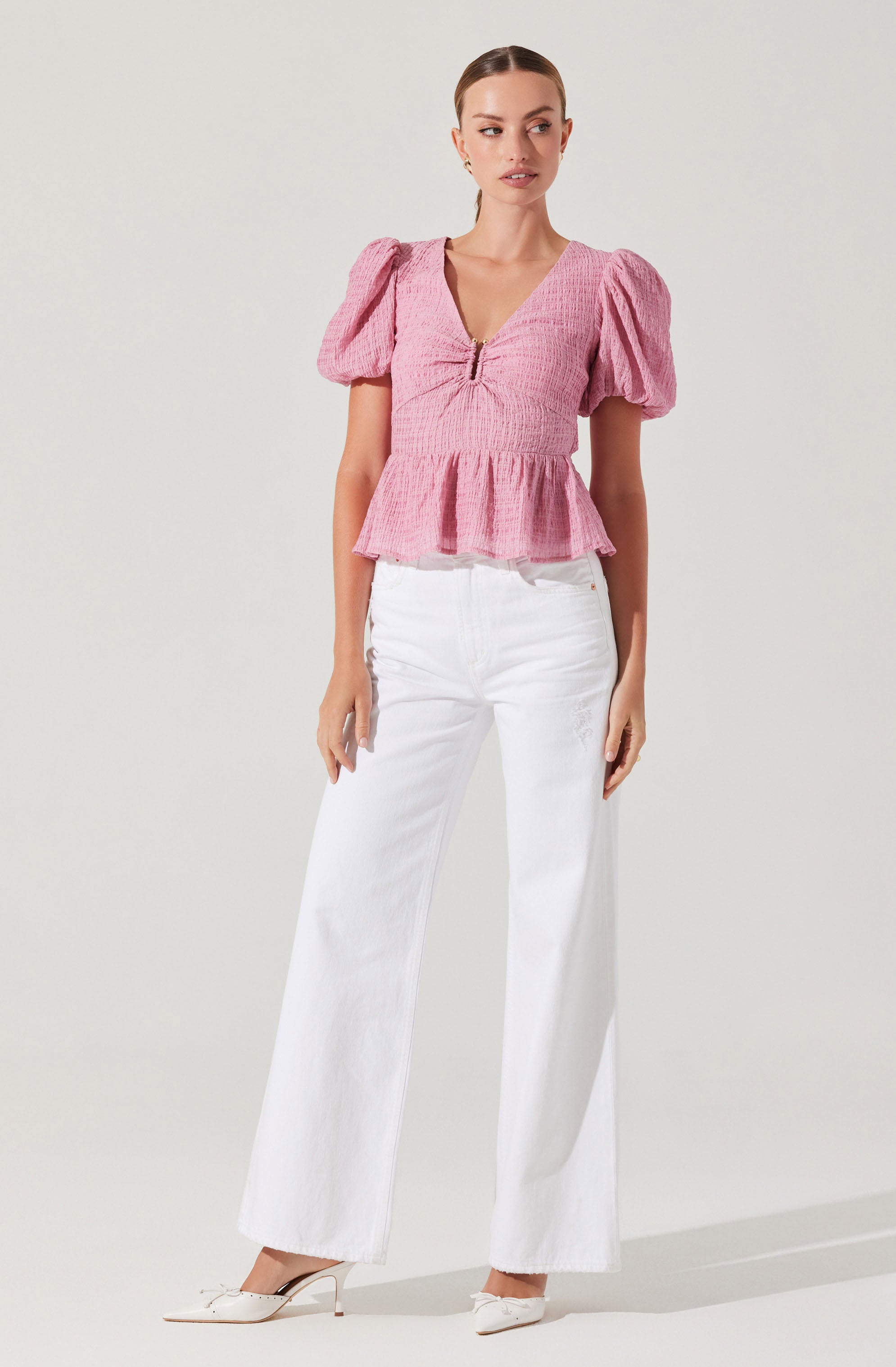 A woman is wearing a pink textured top with puffed short sleeves, a ruched front with a zipper detail, and a peplum hem. She pairs it with high-waisted, wide-leg white jeans and white heeled shoes.