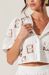 A woman is wearing a white, short-sleeved, button-up top with intricate lace and crochet floral patterns. The top features square cutouts, revealing the skin underneath, and has a slightly cropped length. She is also wearing a gold ring on her finger.