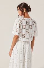 A woman is wearing a matching white lace outfit with a short-sleeved, cropped top and a high-waisted, floor-length skirt. The lace fabric features intricate square and floral embroidery patterns. She accessorizes with gold geometric earrings and has her dark hair styled in a low, twisted bun.