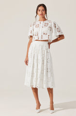 A woman is wearing a white cropped top and a matching white midi skirt, both made from fabric with a geometric cut-out pattern. The top has short sleeves and buttons down the front, while the skirt is high-waisted with a relaxed fit. She is also wearing white pointed-toe shoes.
