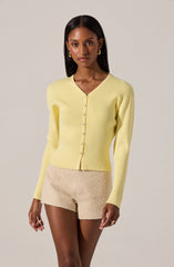 A woman is wearing a light yellow, long-sleeve, V-neck cardigan with matching buttons down the front. She pairs it with beige textured shorts. She has dark, straight hair and is accessorized with gold hoop earrings and a gold ring.