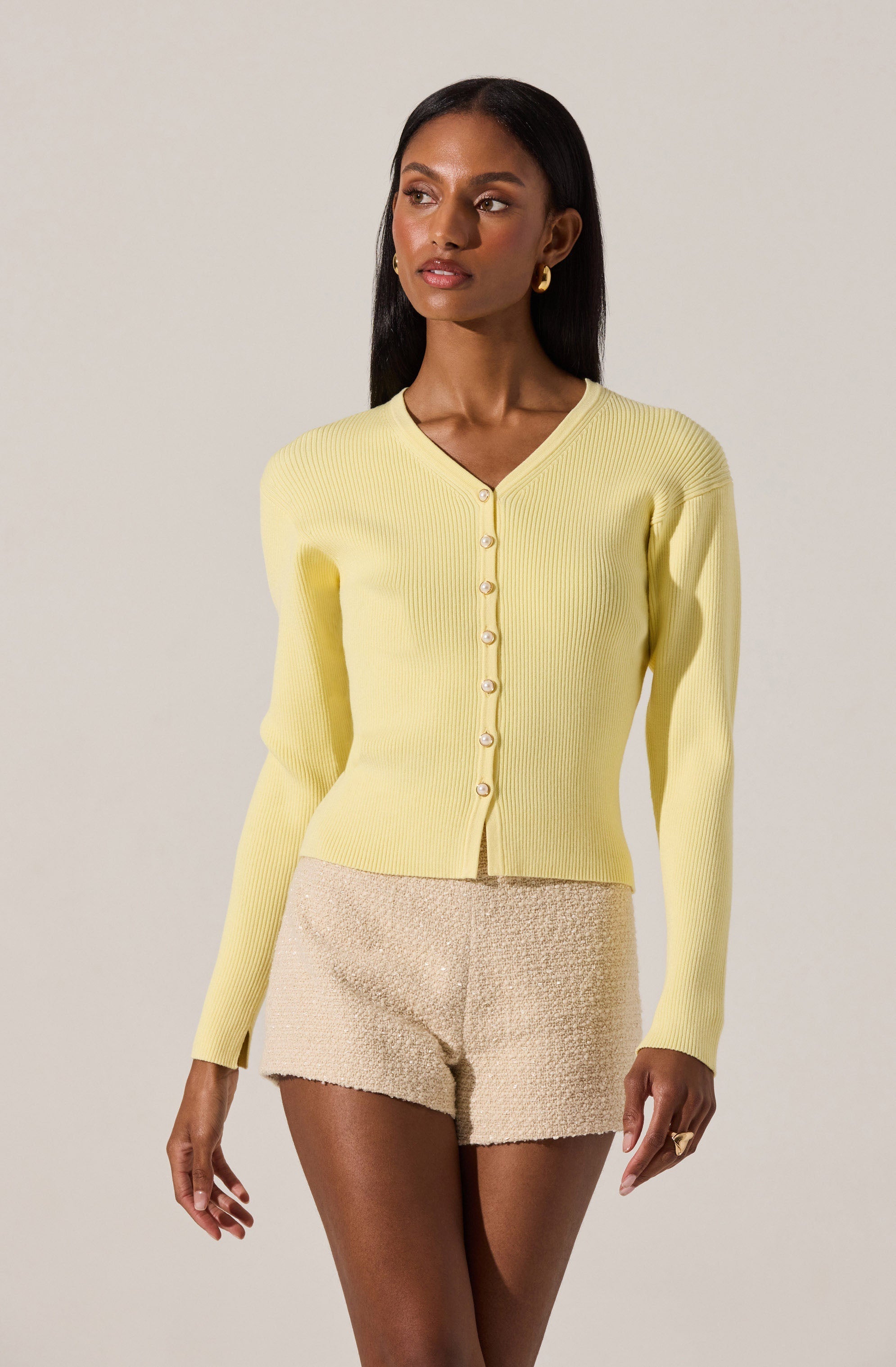 A woman is wearing a light yellow, ribbed, long-sleeve cardigan with a V-neckline and pearl-like buttons down the front. She has paired it with beige textured shorts.