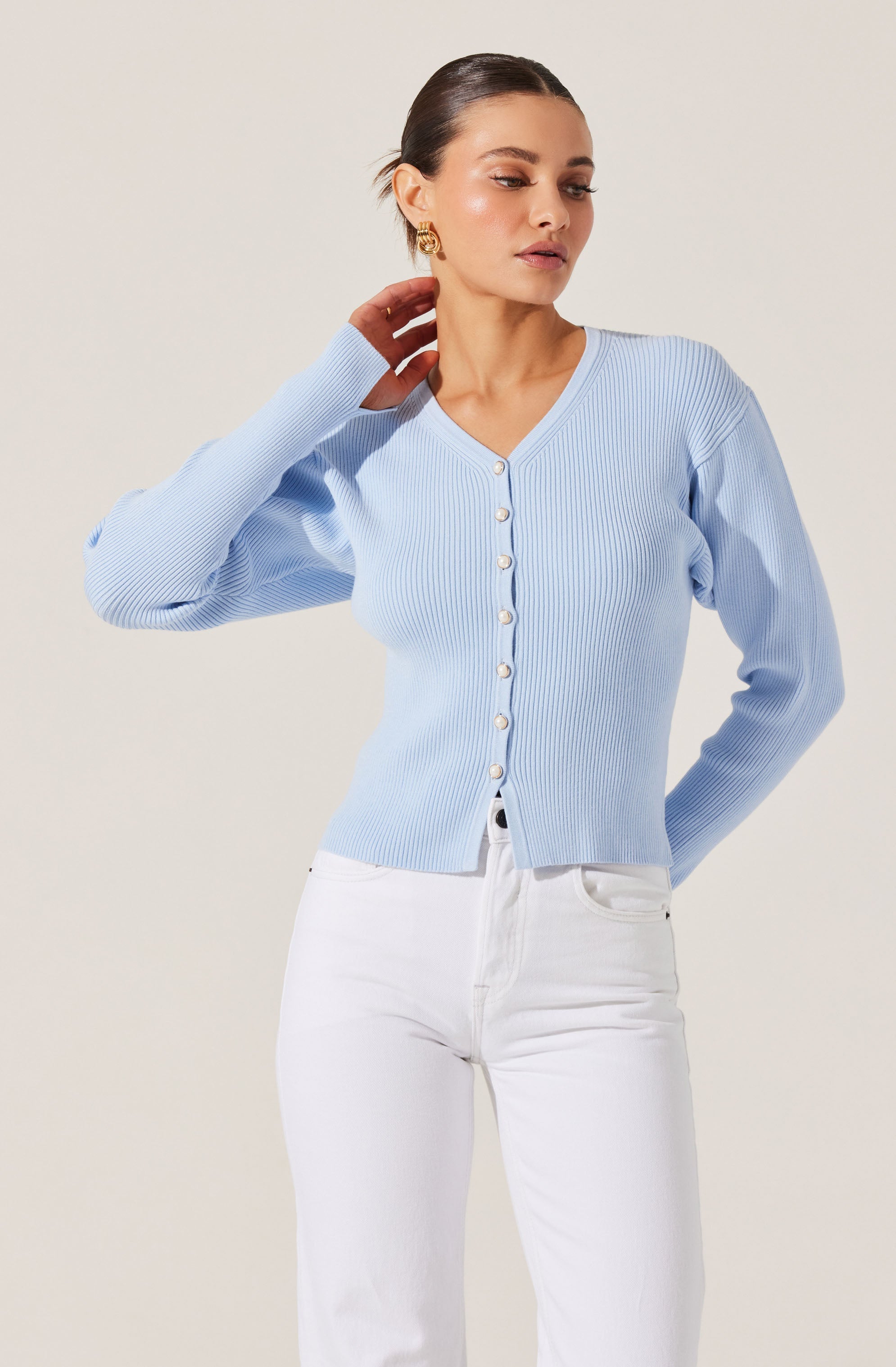 A woman is wearing a light blue, ribbed, V-neck cardigan with long sleeves. The cardigan features a row of white buttons down the front. She has paired it with high-waisted white jeans.