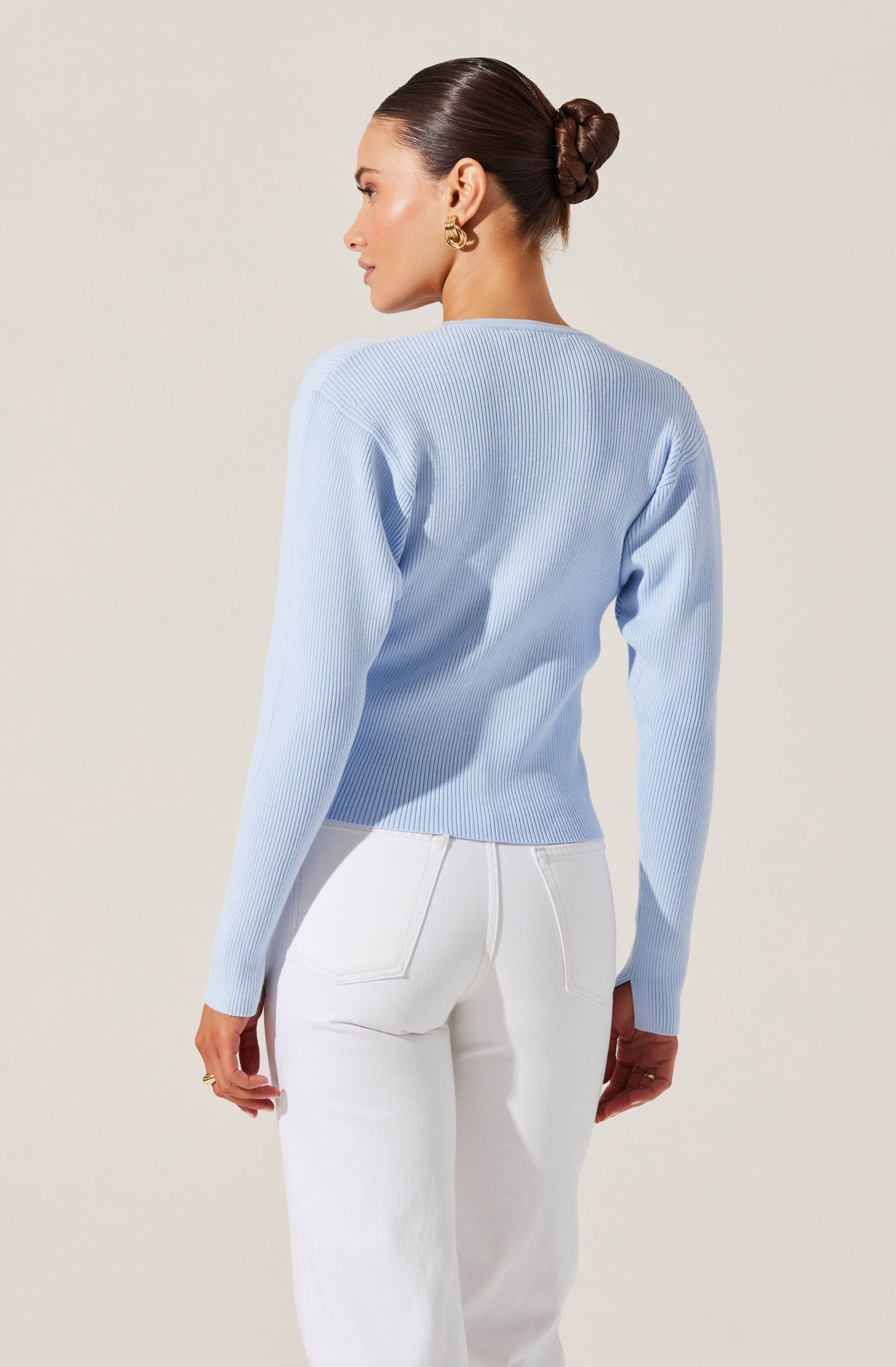 A woman is wearing a light blue, long-sleeved, ribbed sweater with a crew neckline. She is also wearing high-waisted white pants with visible back pockets.
