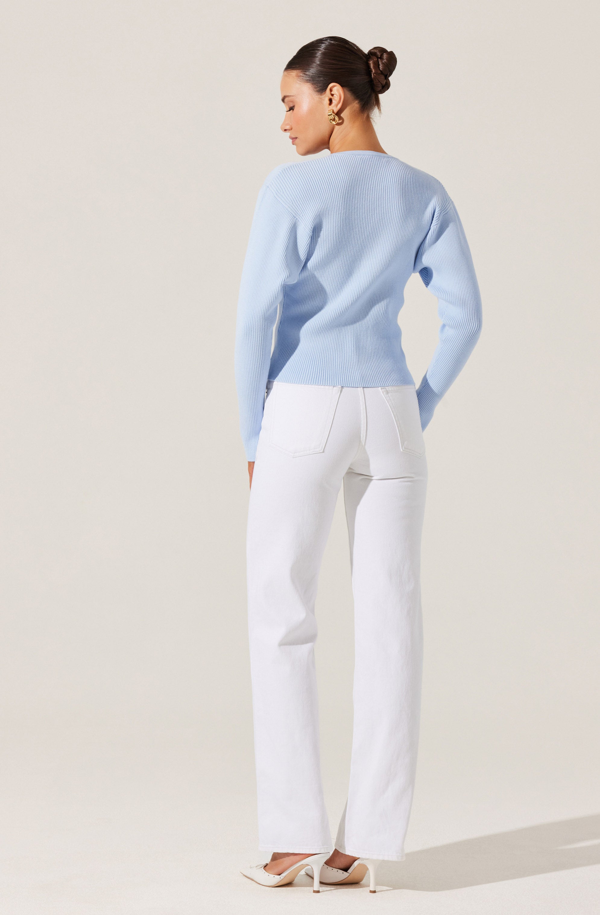 A woman is wearing a light blue, long-sleeve, knit sweater with a relaxed fit. She pairs it with high-waisted, wide-leg white jeans with visible back pockets and a slightly faded wash. She completes the outfit with white high-heeled shoes.