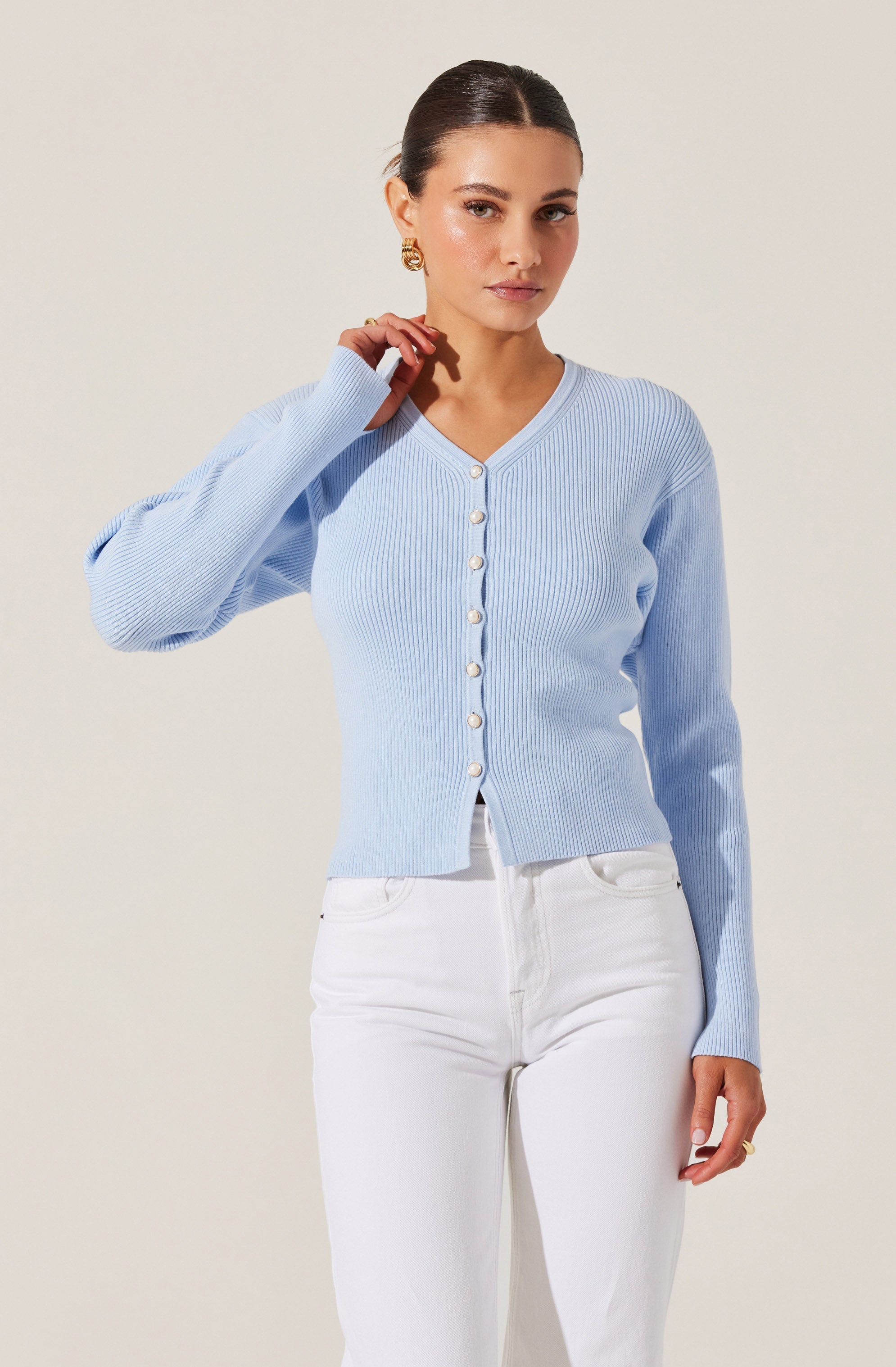 A woman is wearing a light blue, ribbed knit cardigan with a V-neckline and long sleeves. The cardigan features a row of buttons down the front and has a fitted, cropped style. She pairs it with high-waisted white jeans.