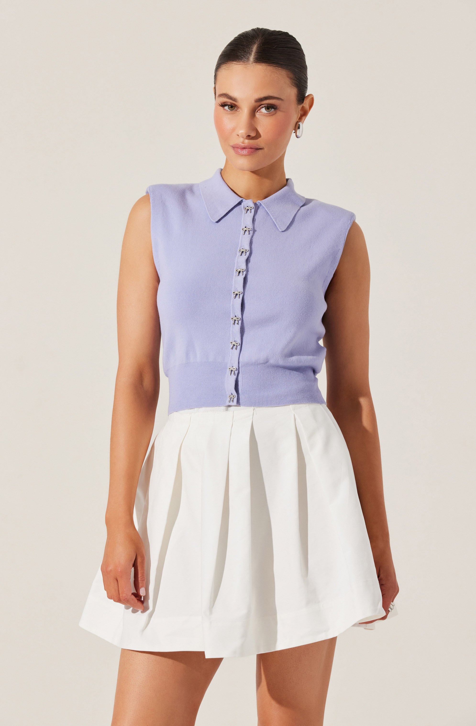 A woman is wearing a sleeveless, light purple, collared knit top with a front button closure, paired with a high-waisted white pleated skirt.