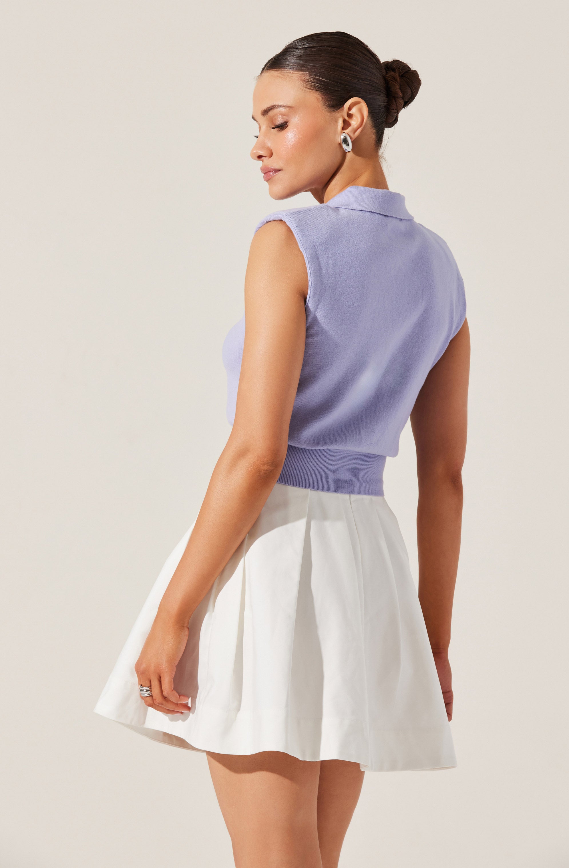 A woman is wearing a sleeveless, lavender-colored top with a collar, made of a soft, textured fabric. She pairs it with a high-waisted, white pleated skirt that reaches above the knees.